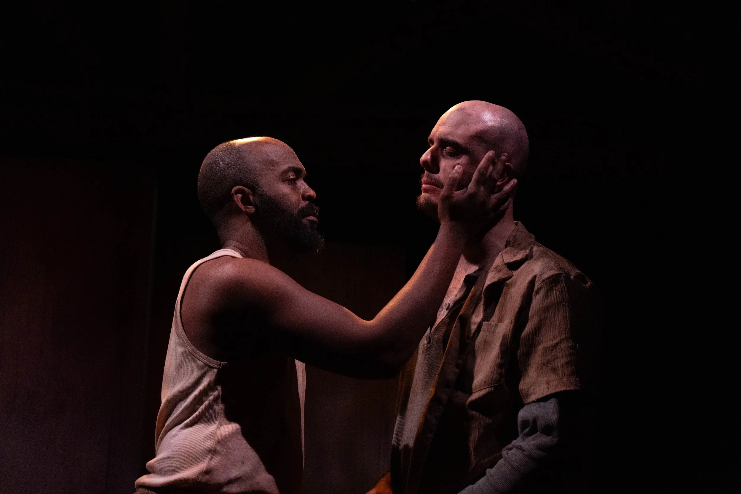 A man with a beard and sleeveless shirt holds the face of a bald man with a beard, who is wearing a brown jacket, as they look at each other against a dark background.