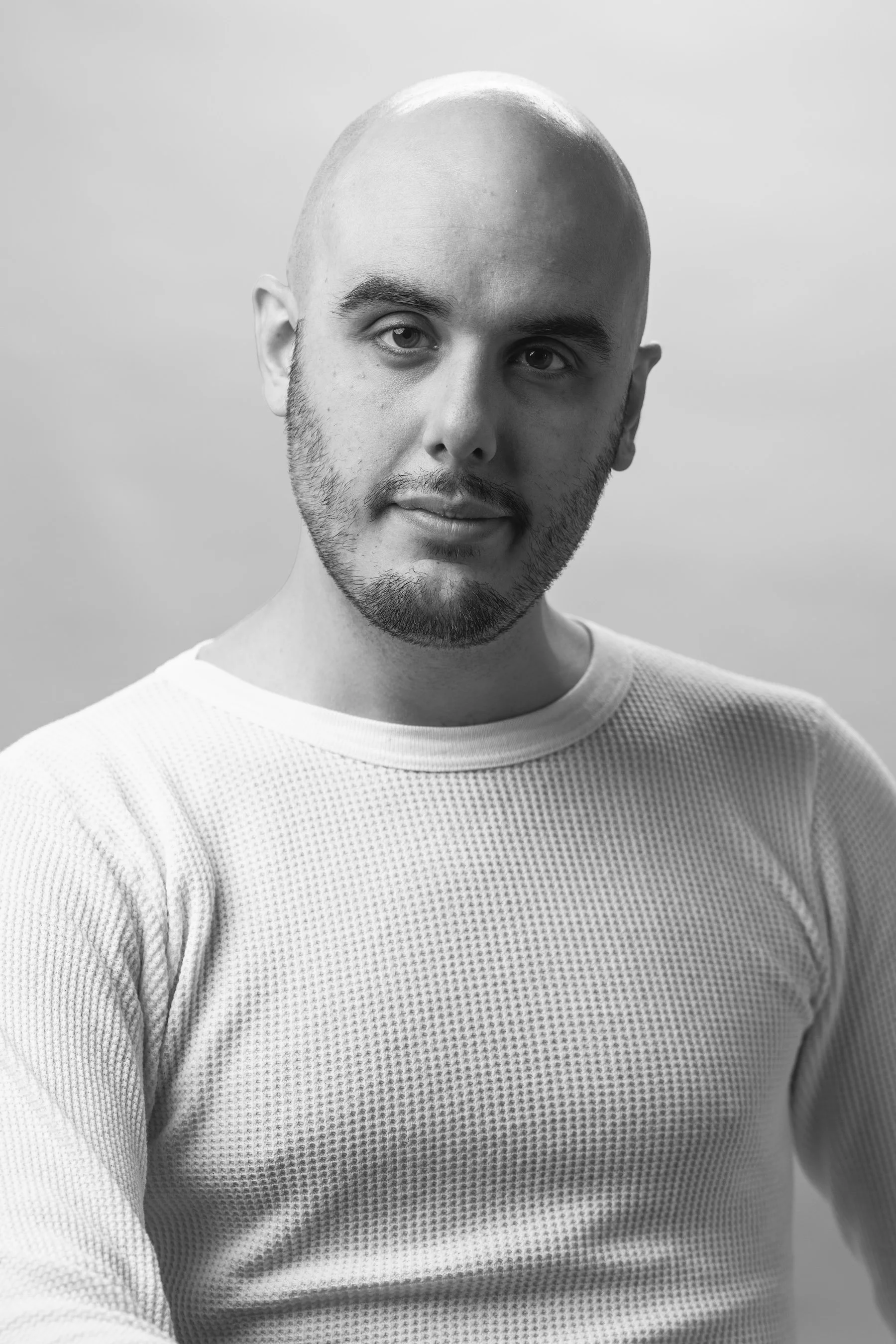 A black and white portrait of a young man with a bald head, beard, and mustache, wearing a textured long-sleeve shirt, against a plain background.