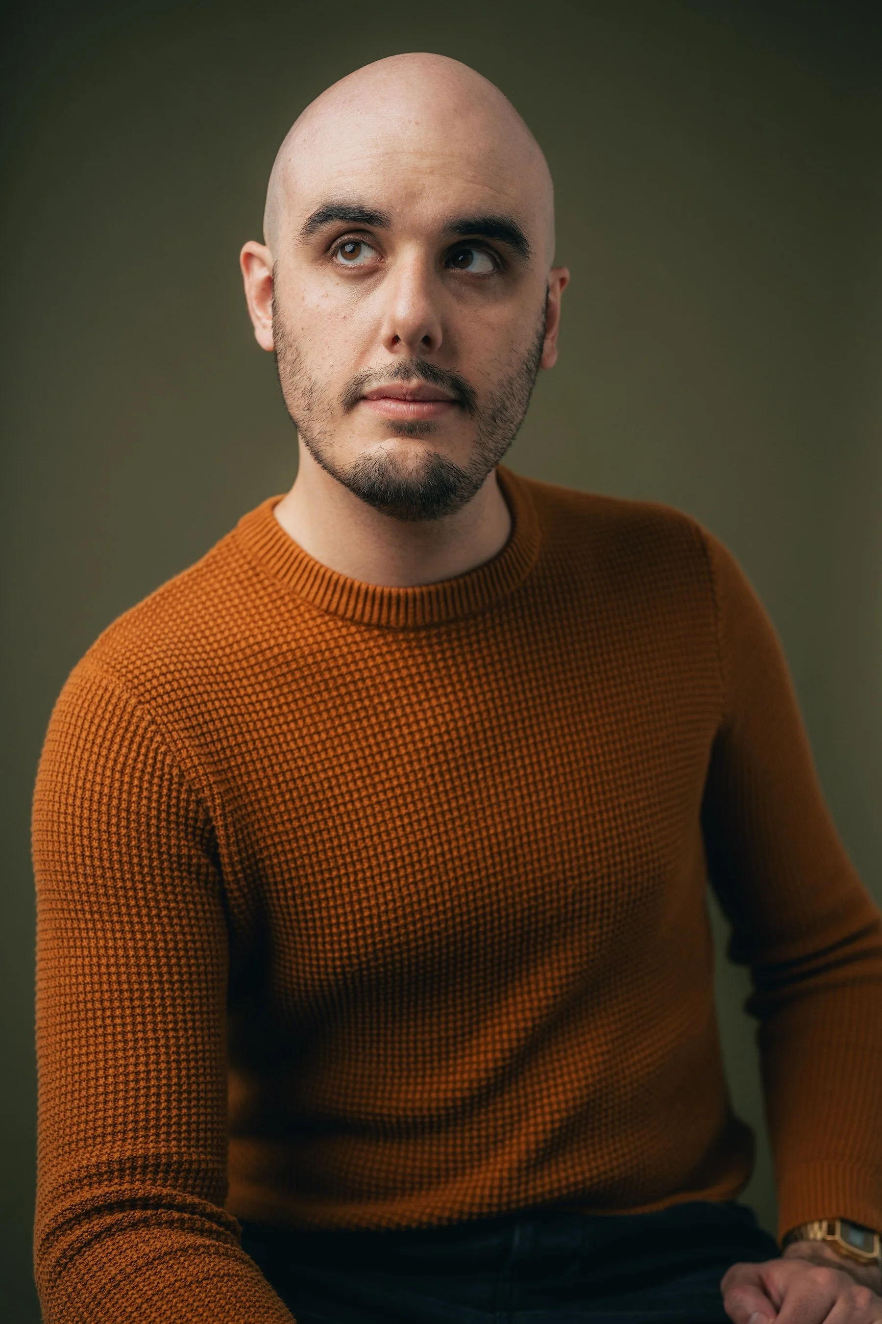 A portrait of a man with a bald head, brown eyes, and facial hair, wearing a textured orange sweater, looking to the side against a greenish background.