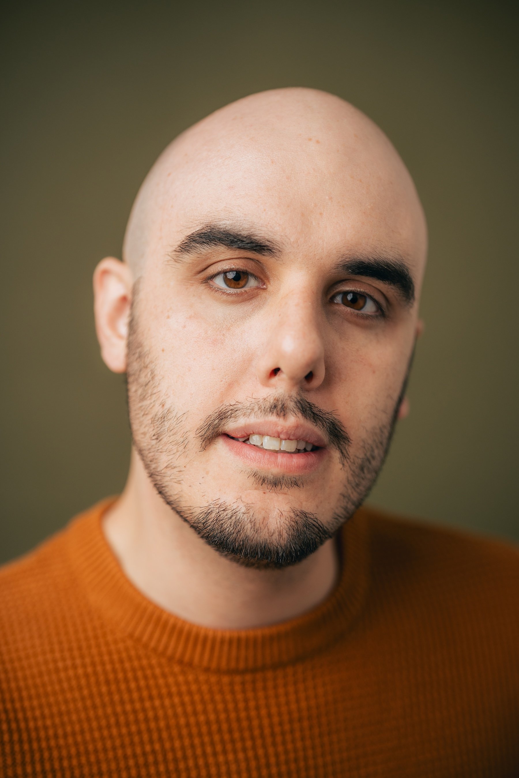 A close-up portrait of a young man with a bald head, brown eyes, and a beard, wearing a burnt orange sweater, looking at the camera with a slight smile.