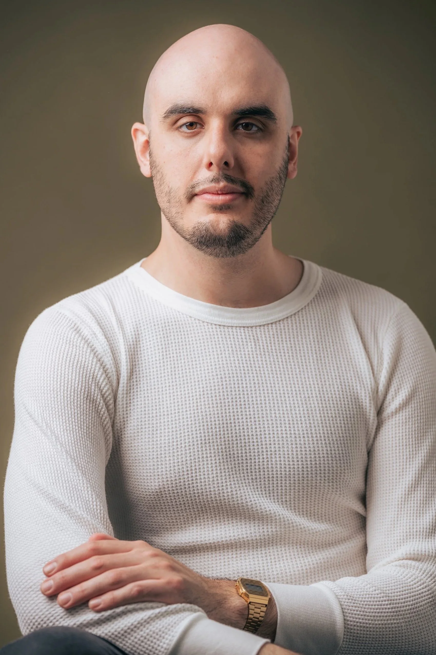 A portrait of a young man with a bald head, beard, and light skin, wearing a white textured long-sleeve shirt and a gold wristwatch, sitting against a brown plain background.