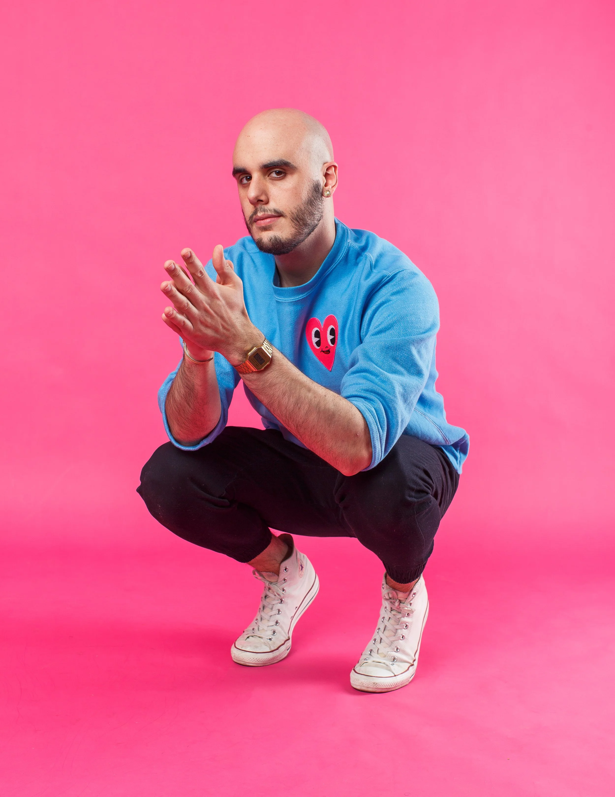 An image of a bald man with a beard wearing a blue sweatshirt with a heart graphic, black pants, and white sneakers, squatting on a neon pink backdrop.