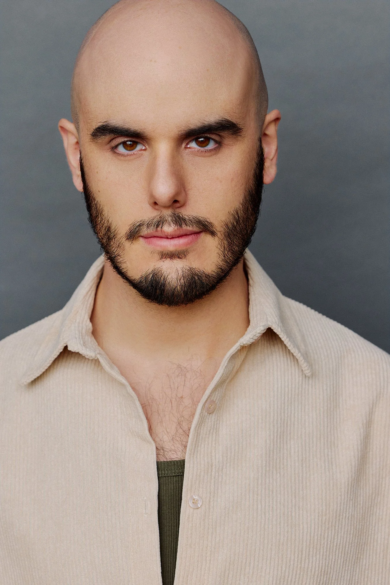 A close-up portrait of a man with a bald head, brown eyes, and a beard, wearing a beige shirt and standing against a charcoal-gray background.