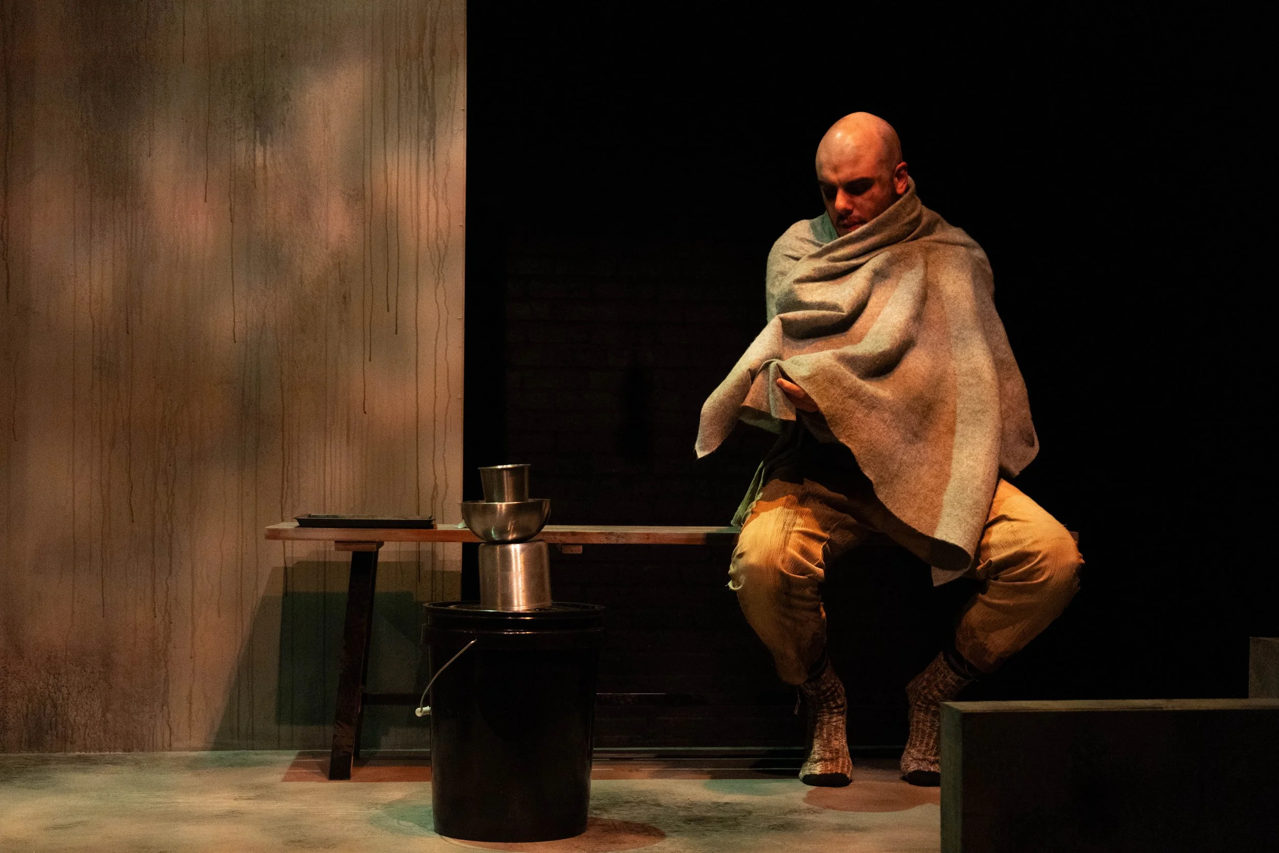 A bald man sitting on a bench, wrapped in a blanket in a dimly lit room, with a black bucket next to him that's balancing a stainless steel drinking cup, a stainless steel bowl, and a pint cup.