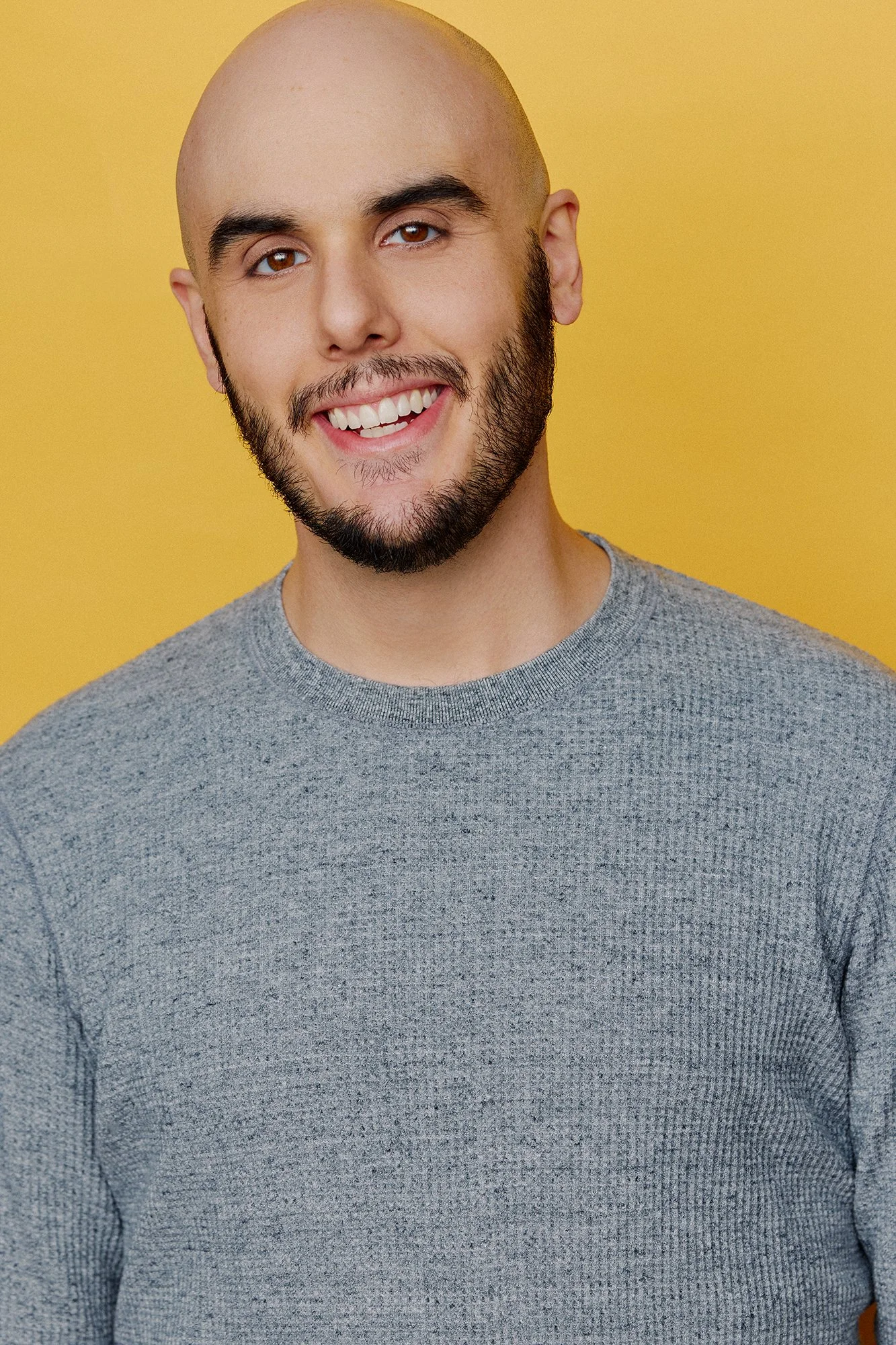 A smiling man with a beard, bald head, and brown eyes, wearing a gray textured long-sleeve shirt against a orange-yellow background.
