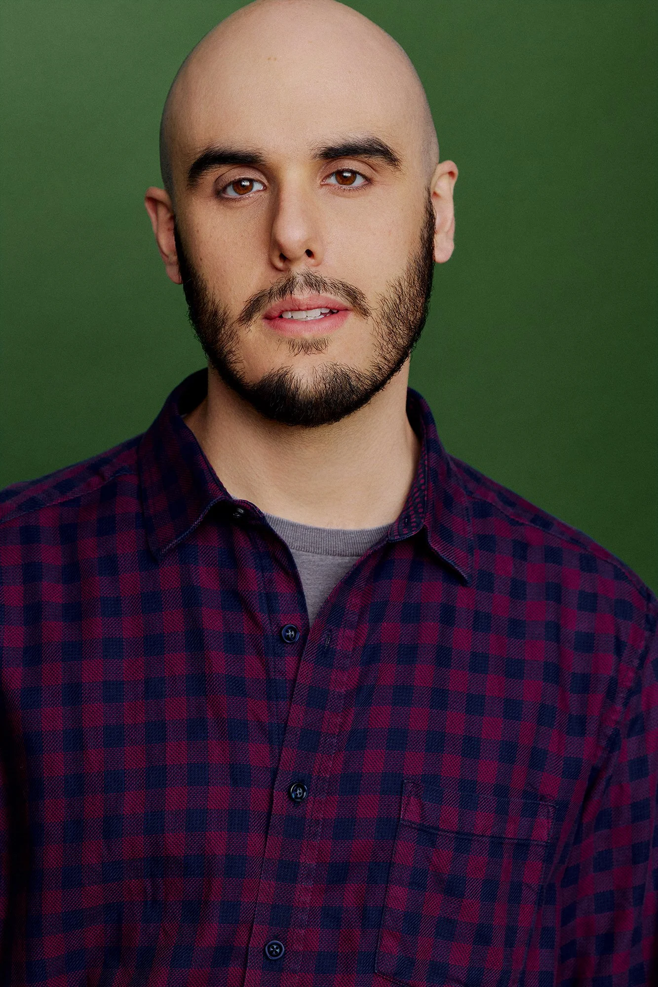 A young man with a bald head and beard wearing a maroon and navy blue checkered shirt against a green background.