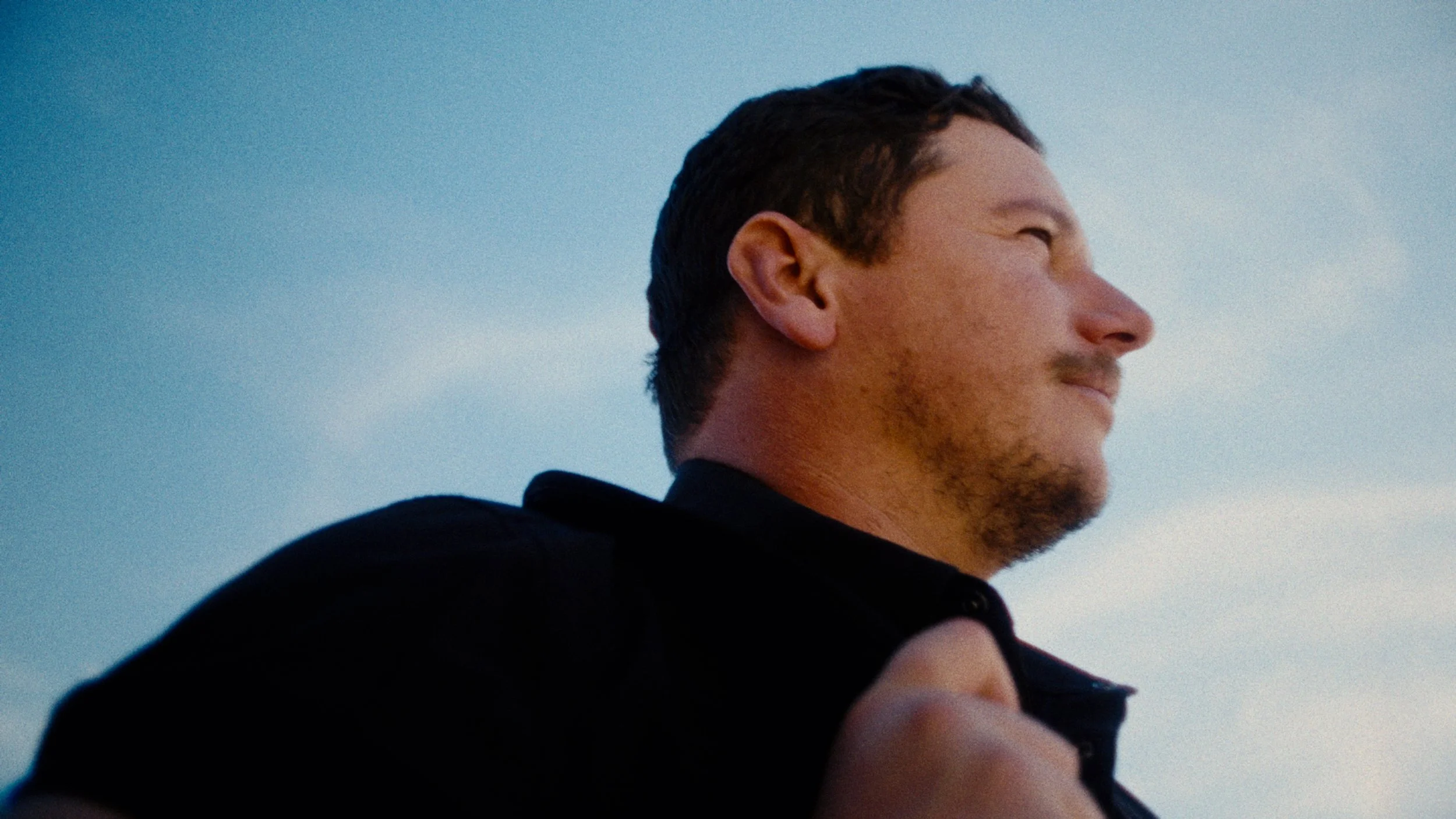 A man with short dark hair looking to the right, holding a strap over his shoulder, against a blue sky background.