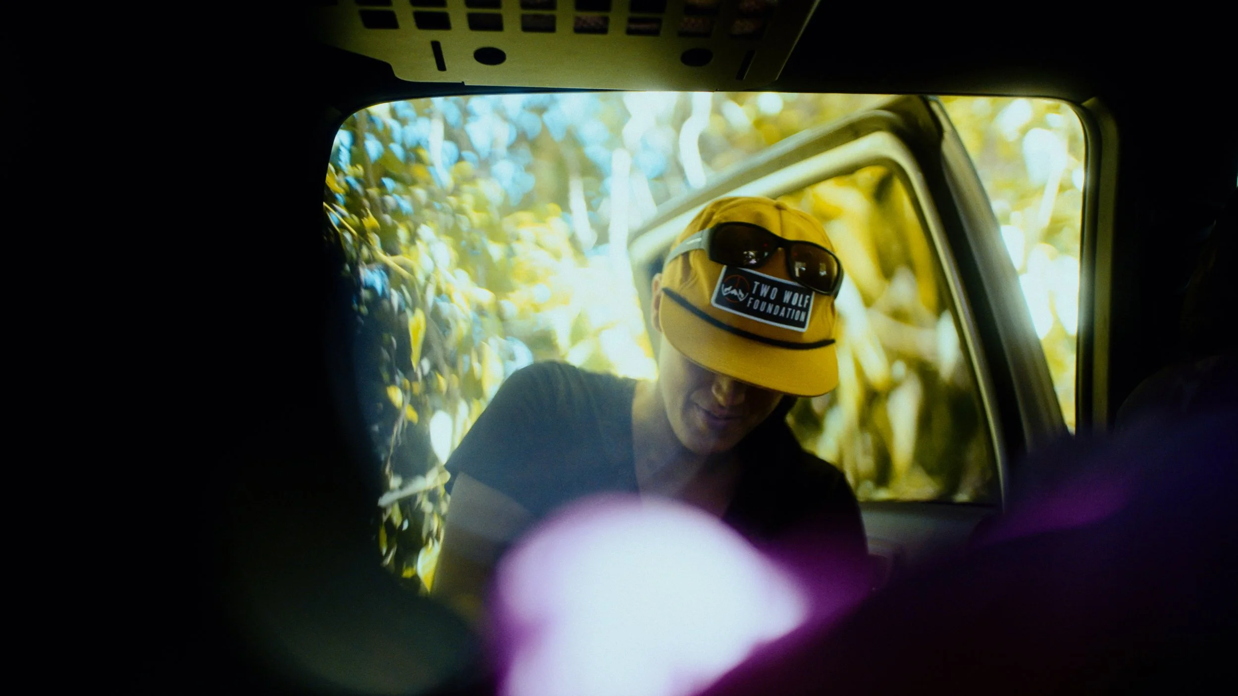 A person wearing a yellow cap with sunglasses and a 'Two Wolf Foundation' sticker, leaning out of a vehicle, surrounded by yellow and green foliage.