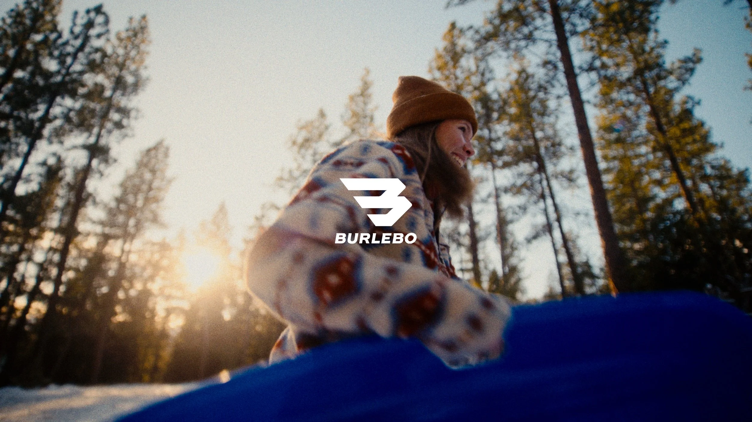 A woman smiling outdoors in a forest at sunset, wearing a patterned jacket and a brown beanie, with a Burlebo logo overlay.