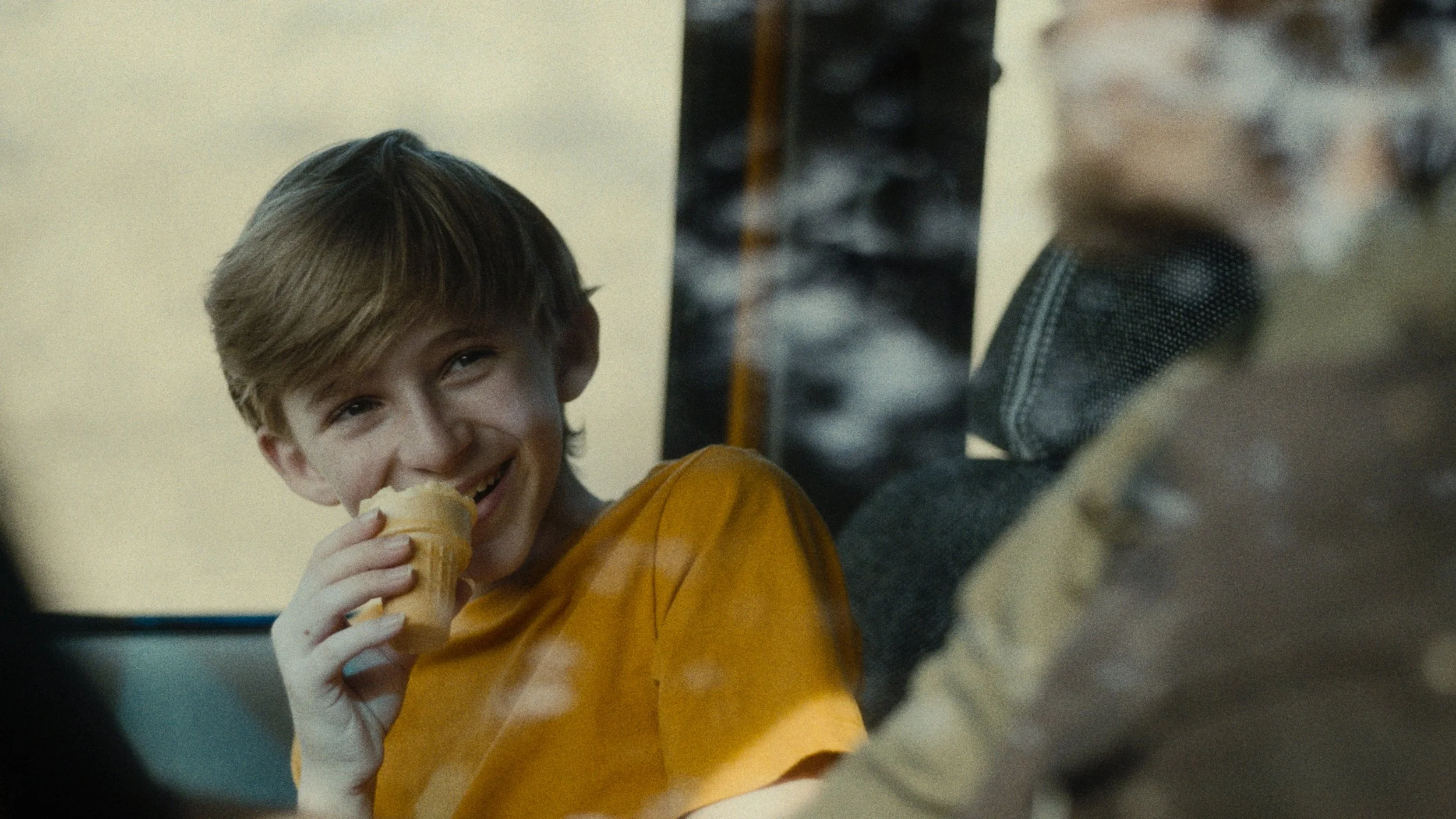 A young boy with light brown hair and a bright smile, wearing a yellow shirt, is holding and eating an ice cream cone. He appears happy and relaxed, sitting indoors with a blurred background.
