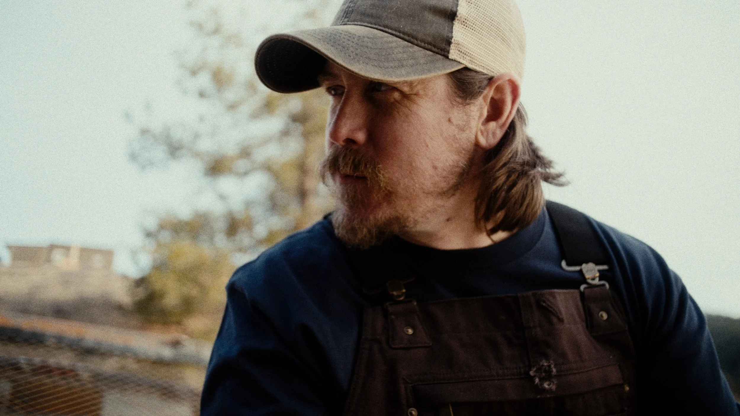 A man with long hair, beard, and mustache wearing a baseball cap and a dark apron.