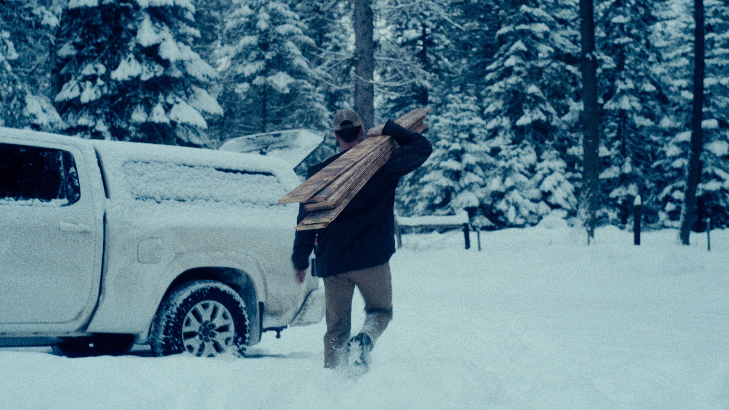 A person walking through snow carrying a stack of wooden planks near a white pickup truck in a snow-covered forest.
