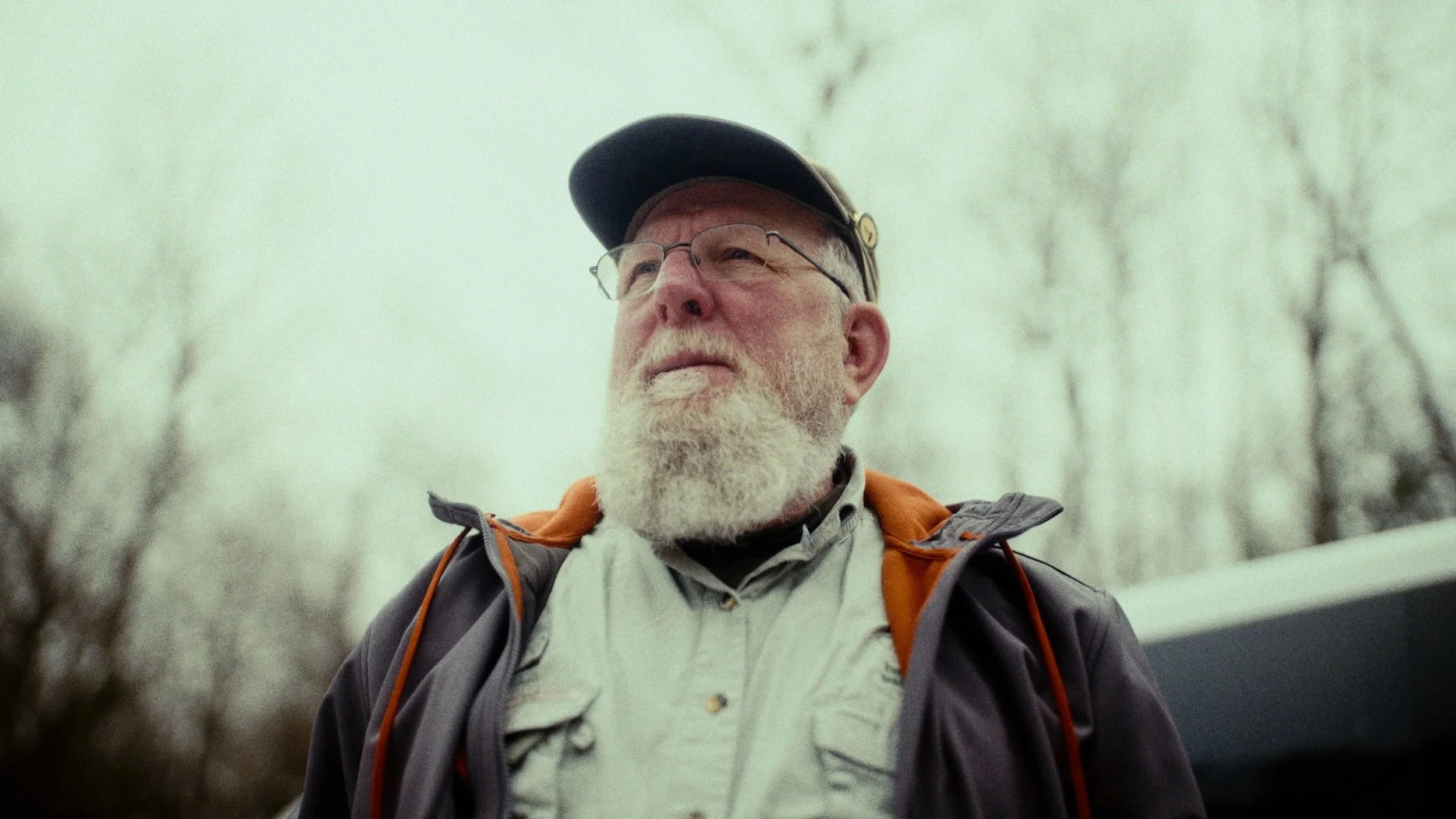 An older man with a white beard and glasses, wearing a black cap and outdoor jacket, outdoors in a forested area with bare trees.