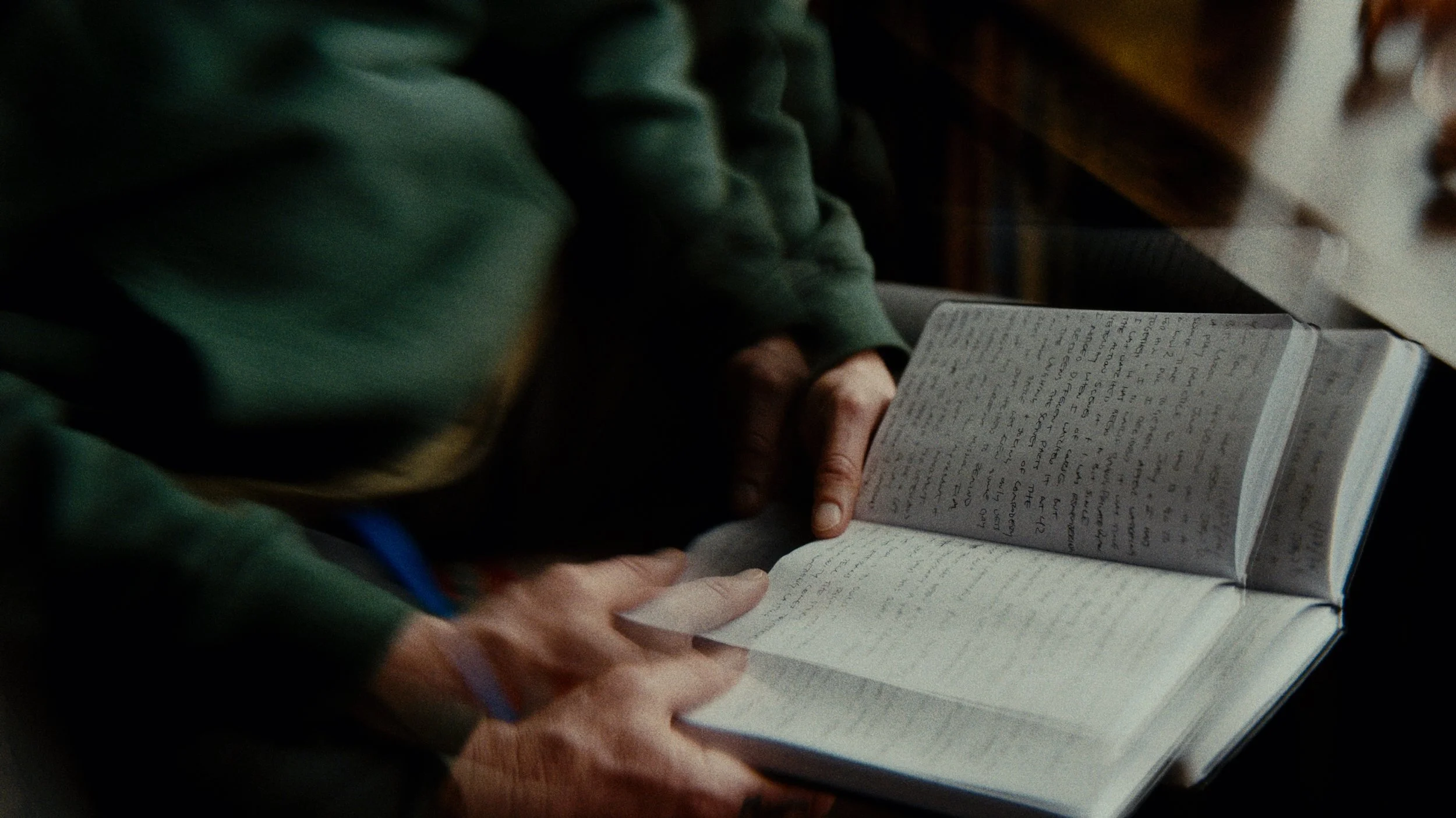 A person wearing a dark green hoodie reading a handwritten journal in a dimly lit room.