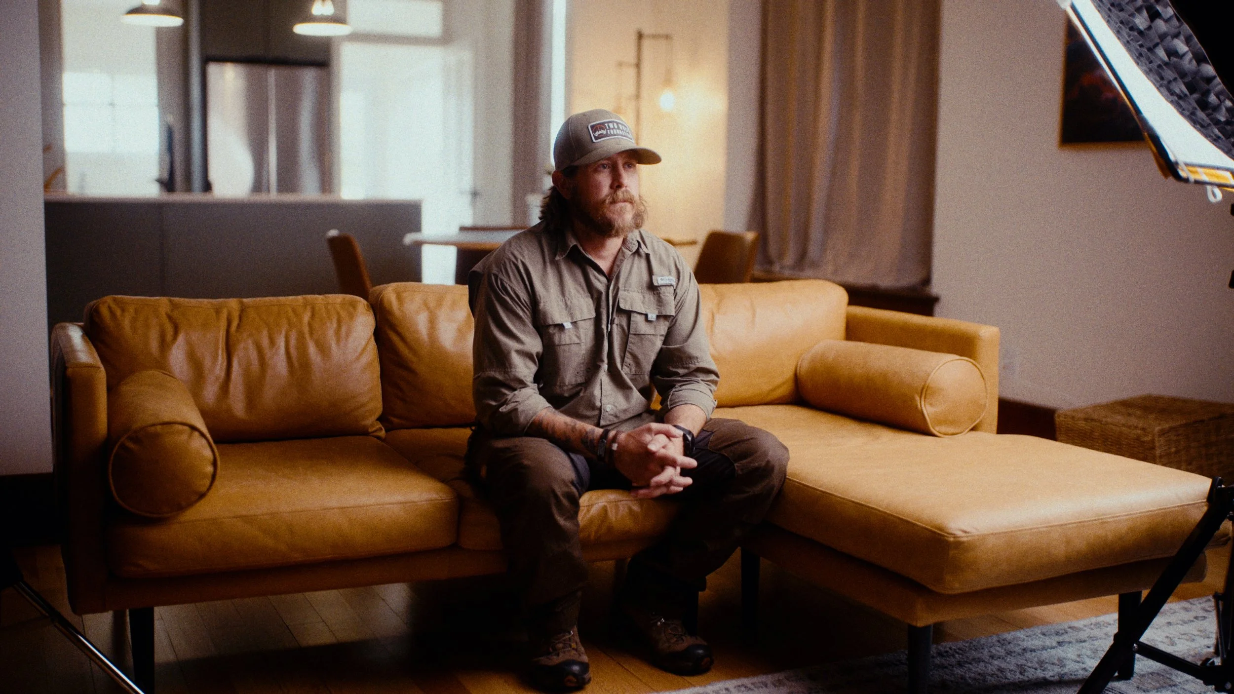 A man sitting on a yellow leather sofa in a warmly lit living room during a photo or video shoot.
