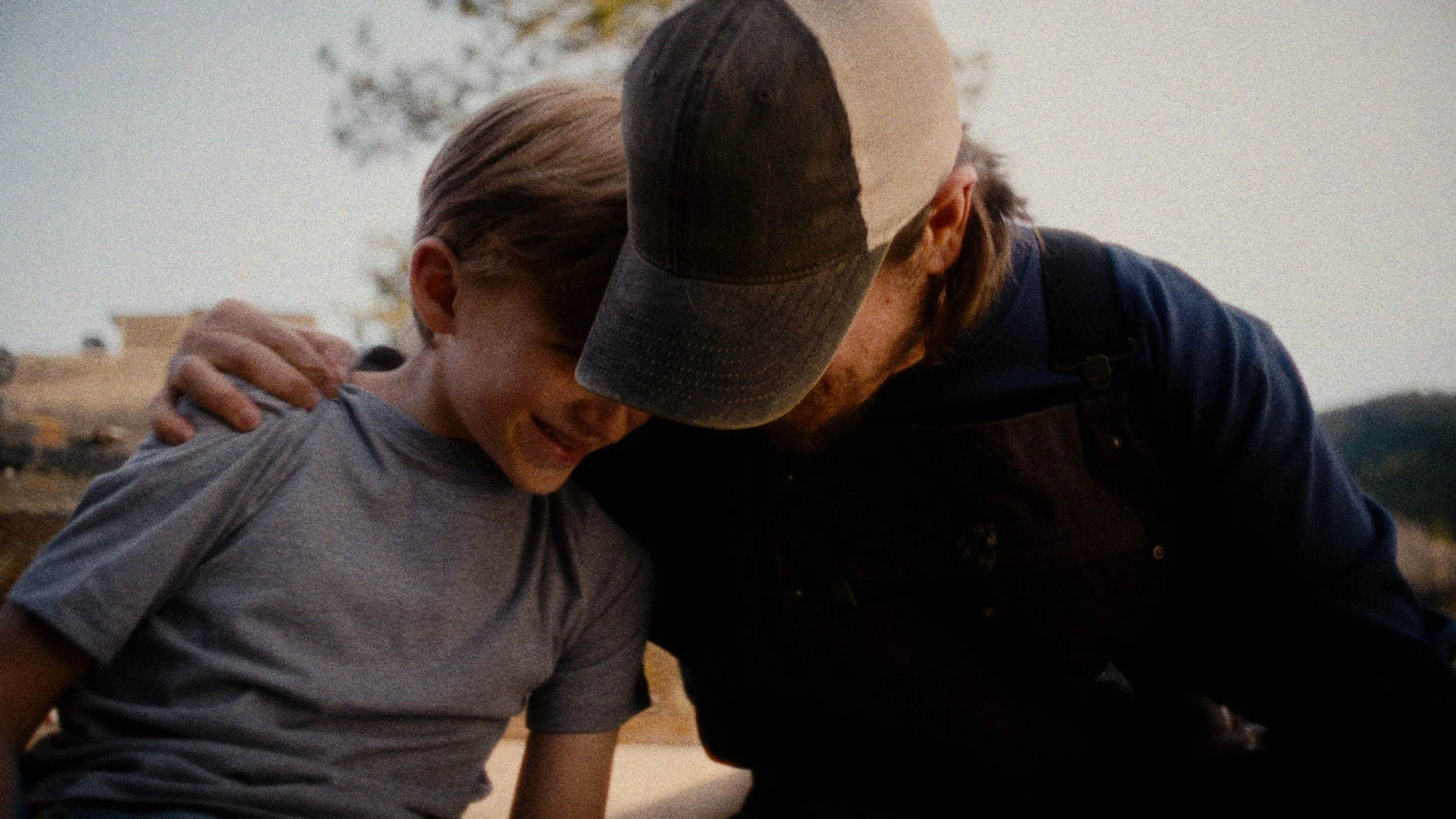 A young boy and a man with a beard share an emotional moment outdoors, their foreheads touching and eyes closed, with the boy smiling.