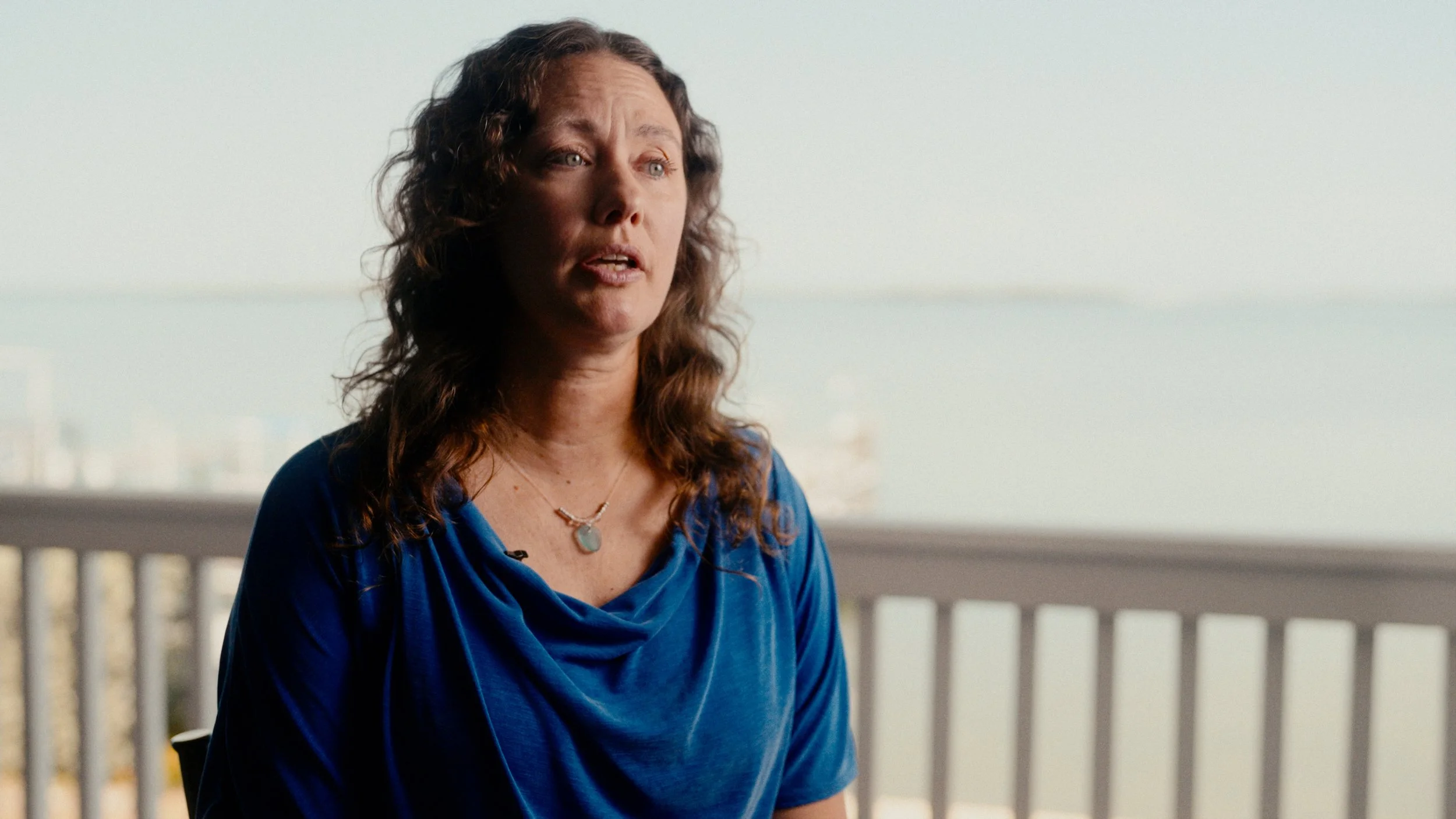 A woman with wavy brown hair and light skin sitting on a balcony, wearing a blue top and a necklace, speaking with a city view in the background.