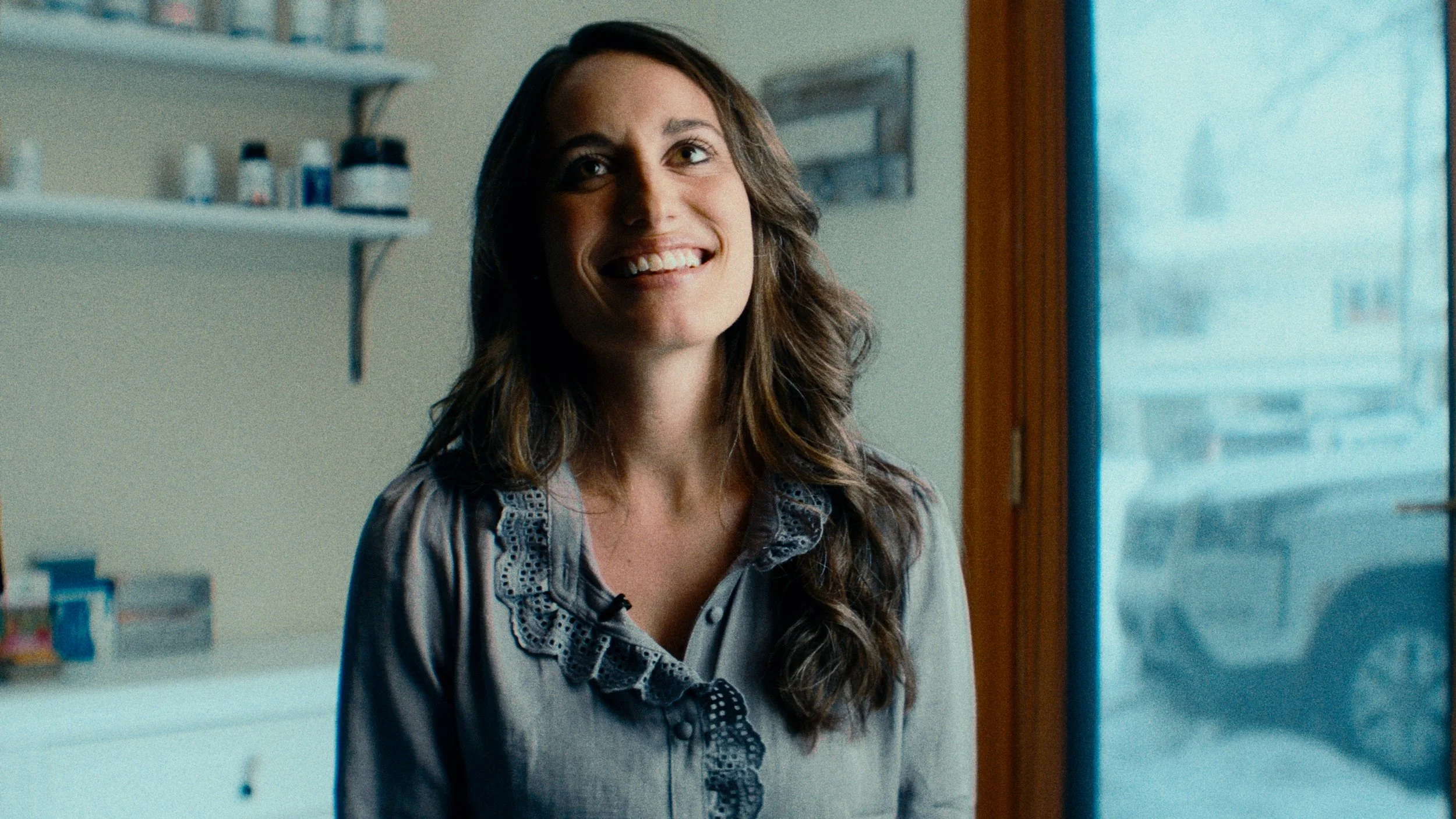 A woman with long brown hair and a grey top smiling inside a room.