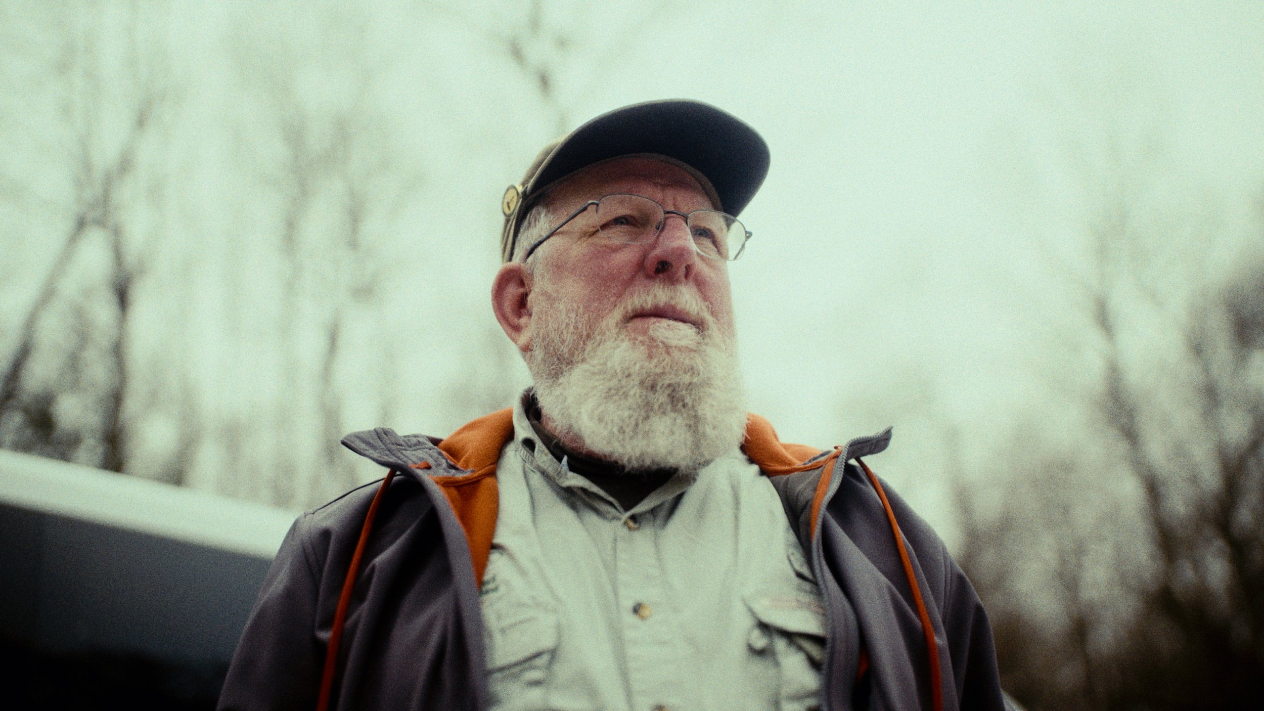 An older man with a white beard and glasses wearing outdoor gear, including a cap and jacket, looking into the distance.
