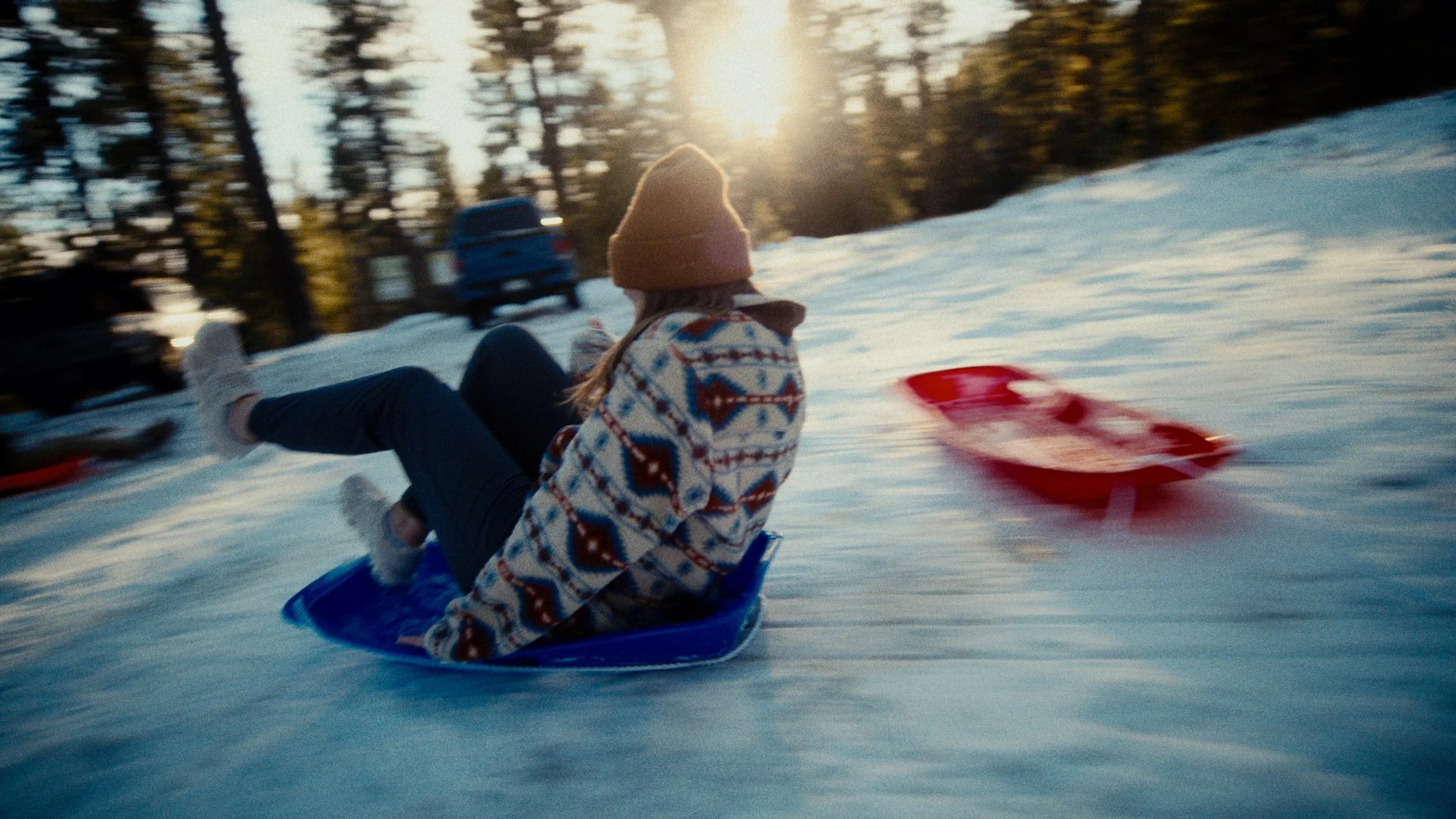 A person in winter clothing, including a colorful sweater and a brown beanie, is sitting on a blue sled moving downhill in the snow during sunset, with trees and parked cars visible in the background.