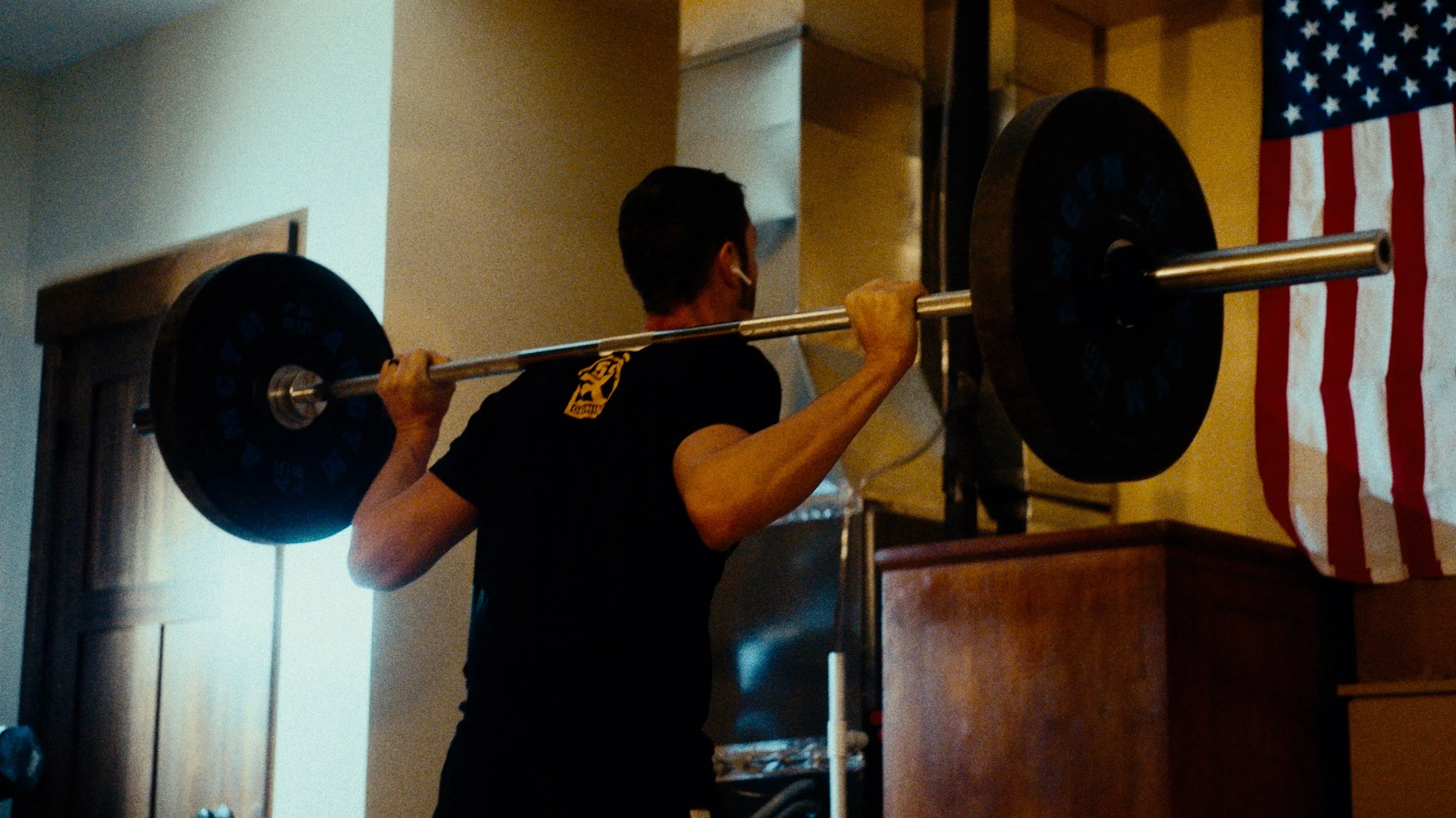 Person lifting a barbell with weights in a gym, American flag in the background.