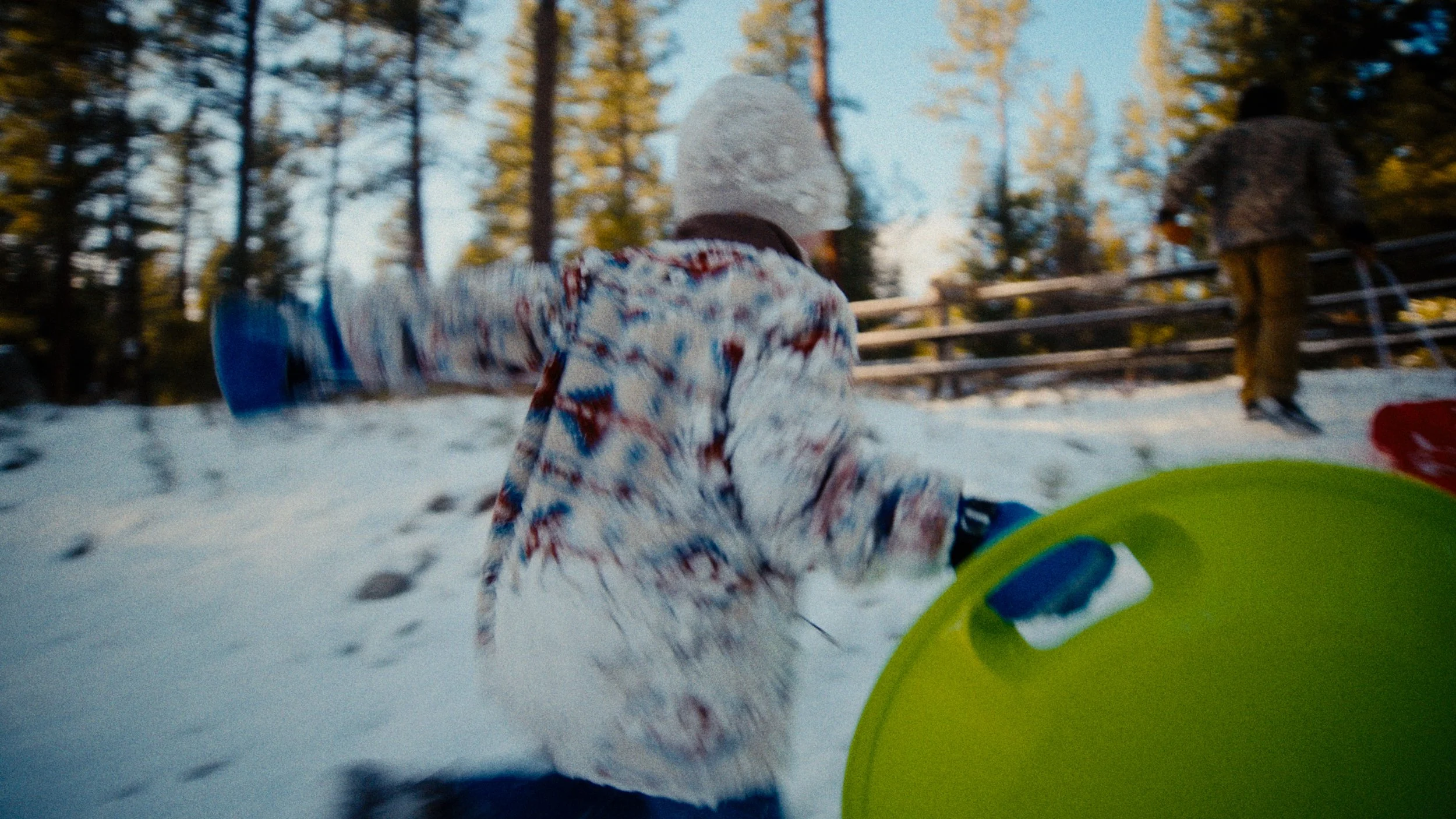 Children sliding down snow-covered hill outdoors during winter, with trees and wooden fence in background.