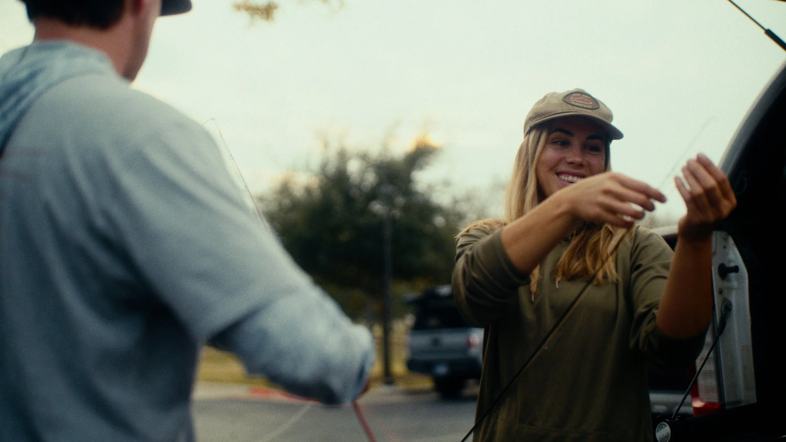 A young woman with long blonde hair and a beige cap smiling as she attaches a wire to a small electronic device, while a man with short dark hair, seen from behind, wears a gray hoodie and holds a wire. They are outdoors near a vehicle with an open t