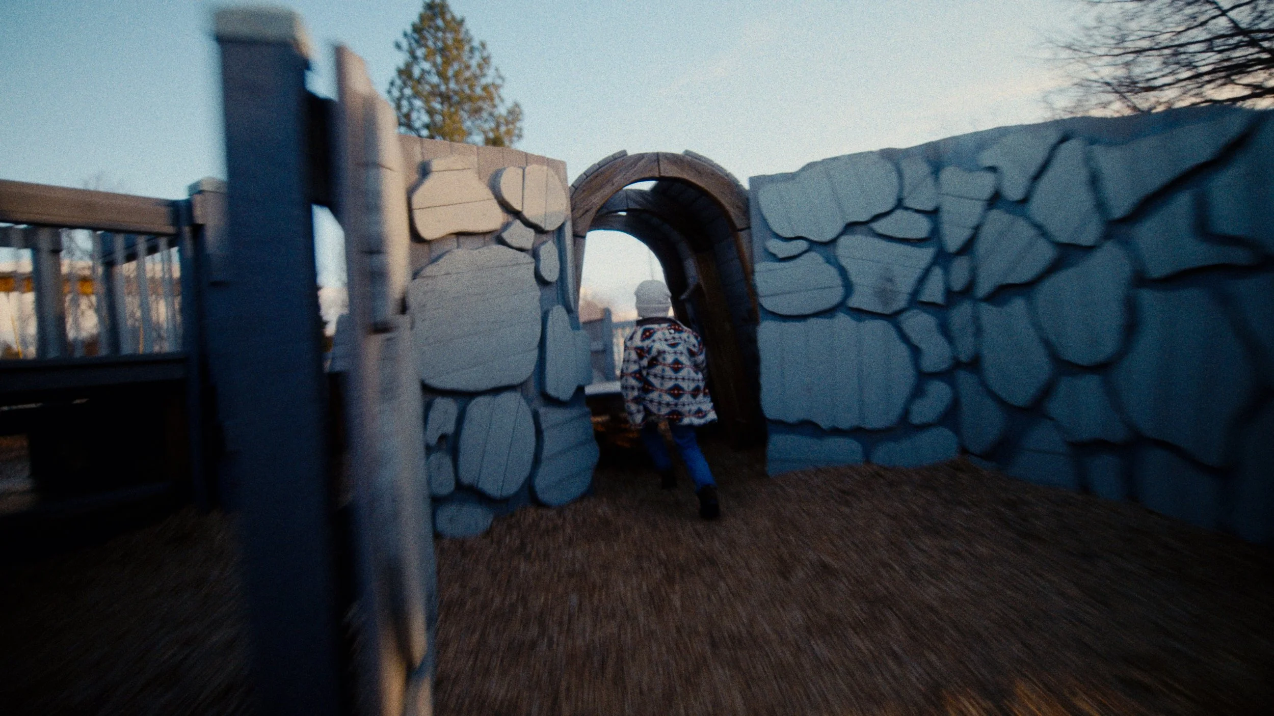 Child wearing a jacket and hat walking through a tunnel made of stone-like structures on a playground during sunset.