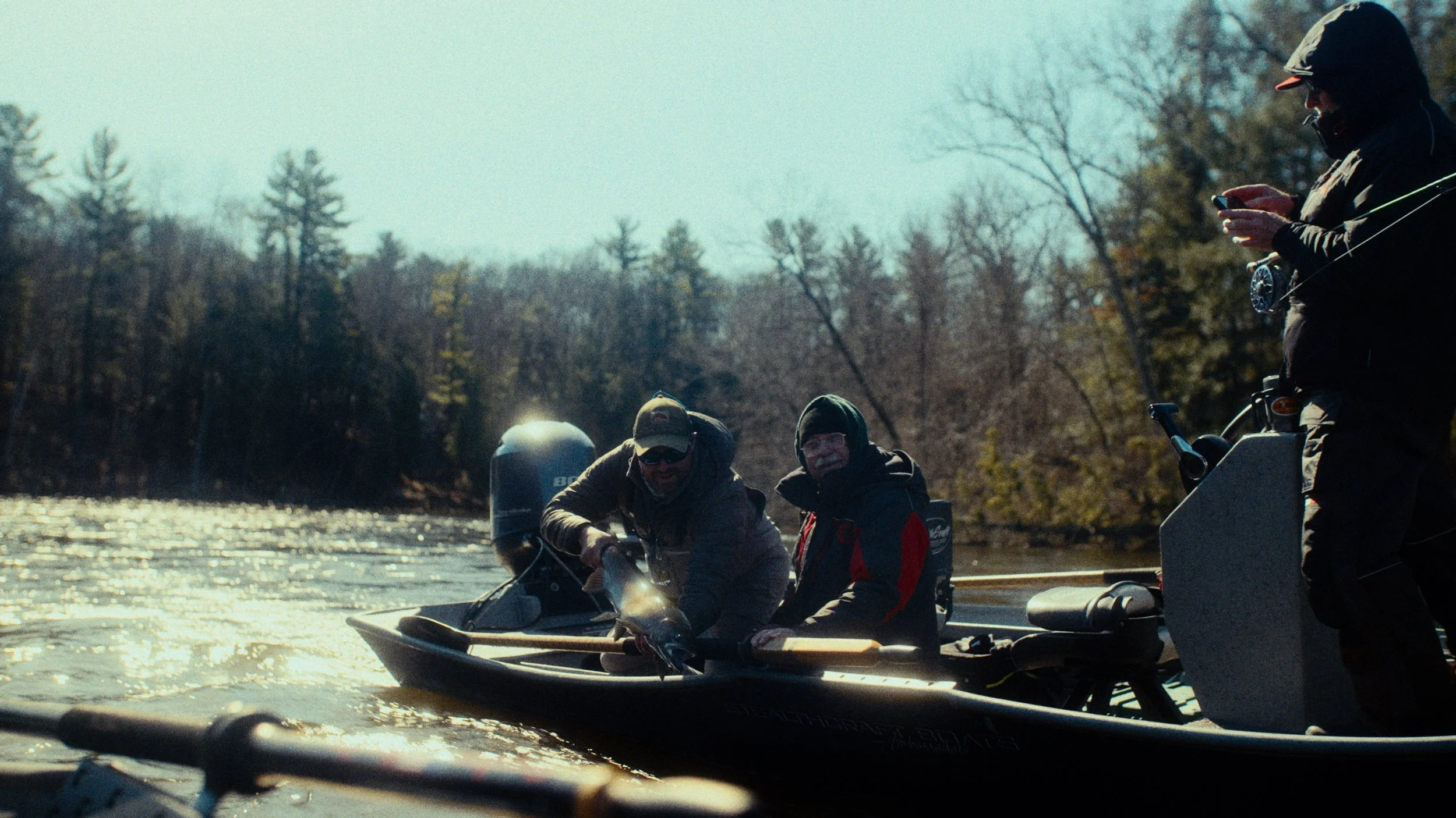 Three people in a boat on a river, with one person working a fishing line and two others watching, surrounded by trees with autumn foliage.