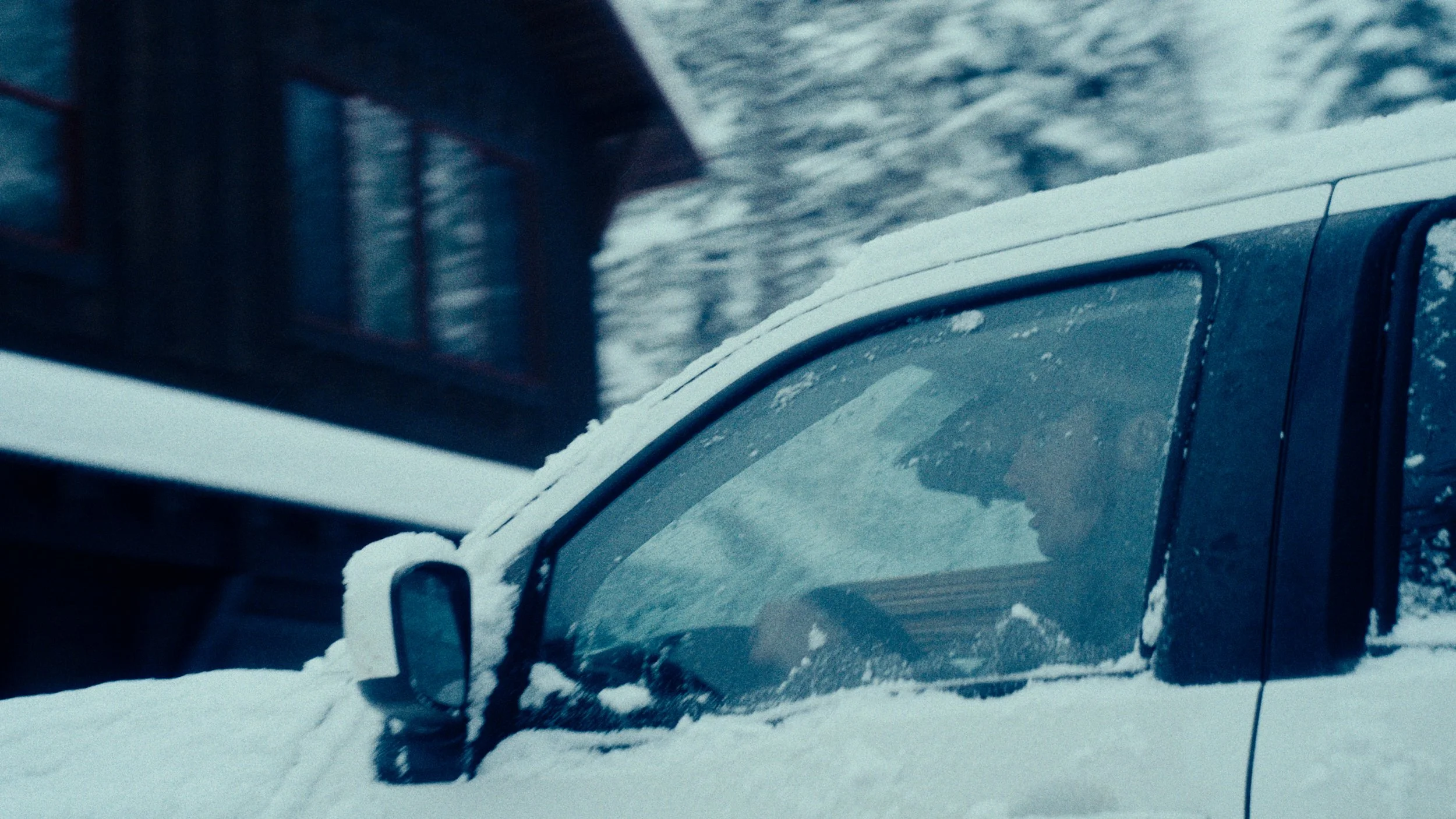 A black vehicle covered in snow, with a person inside wearing sunglasses and a hat, driving through a snowy landscape.