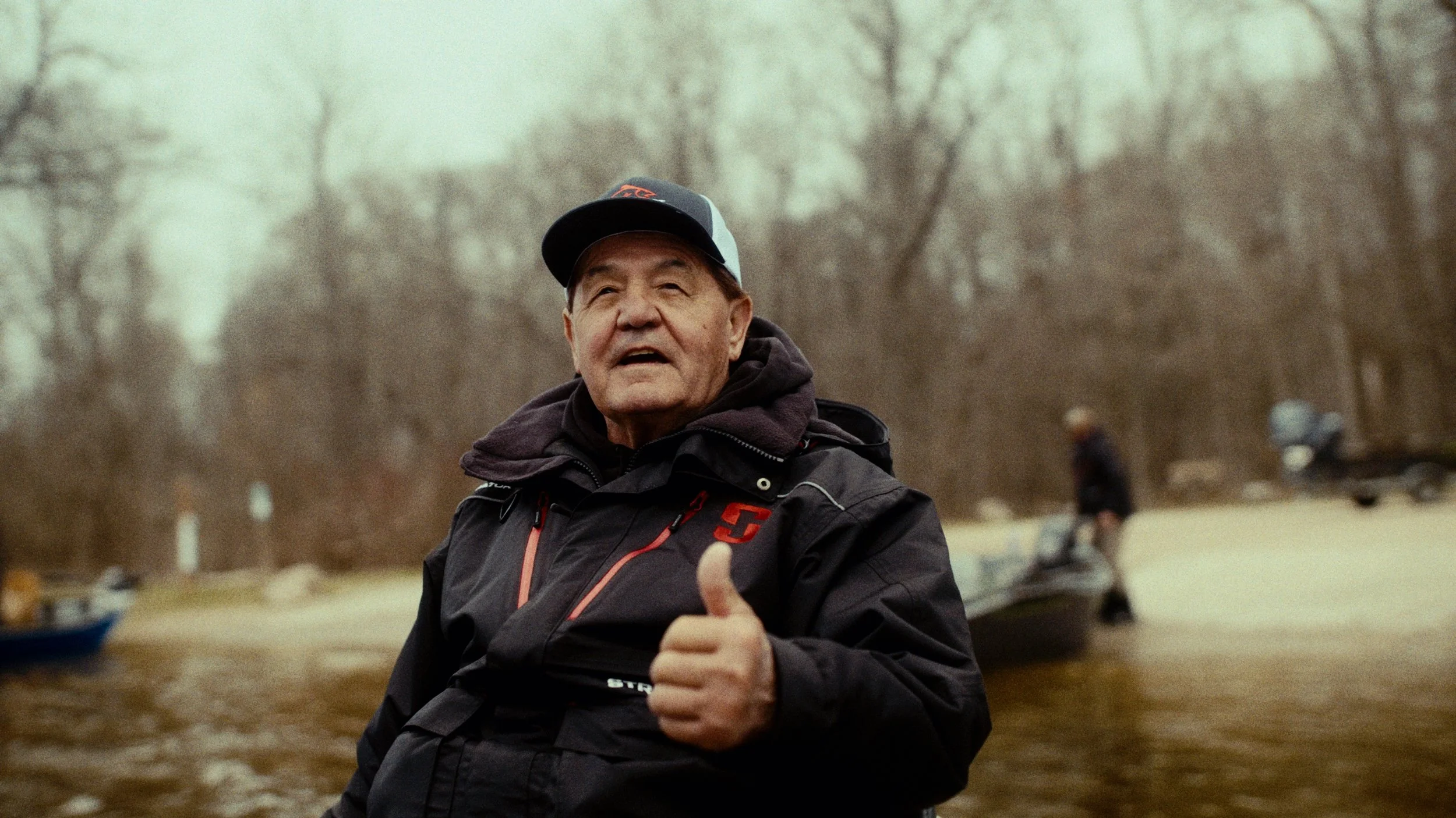 An elderly man wearing a black jacket and a baseball cap giving a thumbs-up gesture outdoors near a body of water with boats in the background.
