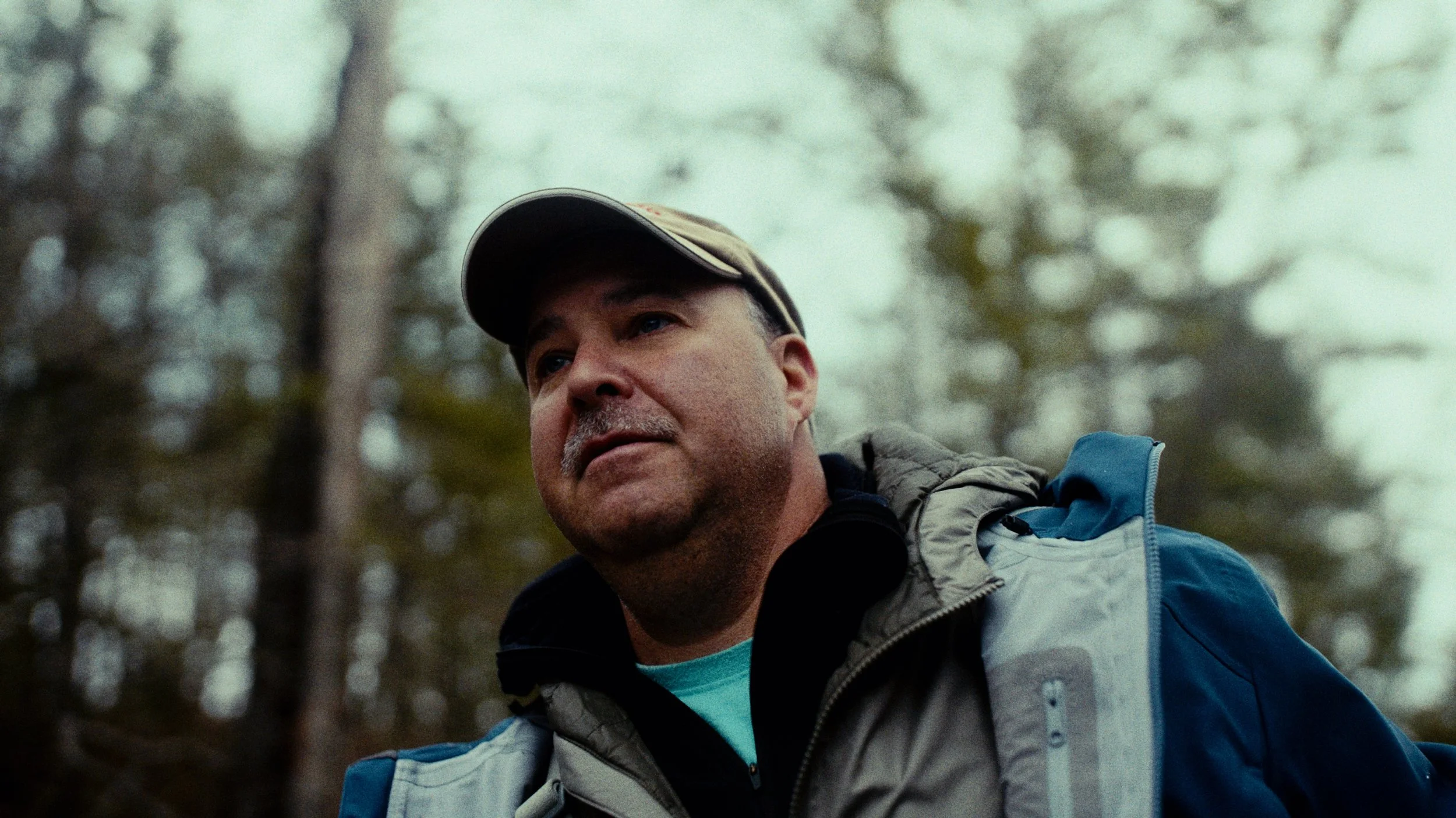 A man outdoors wearing a baseball cap, jacket, and backpack with a forest background.