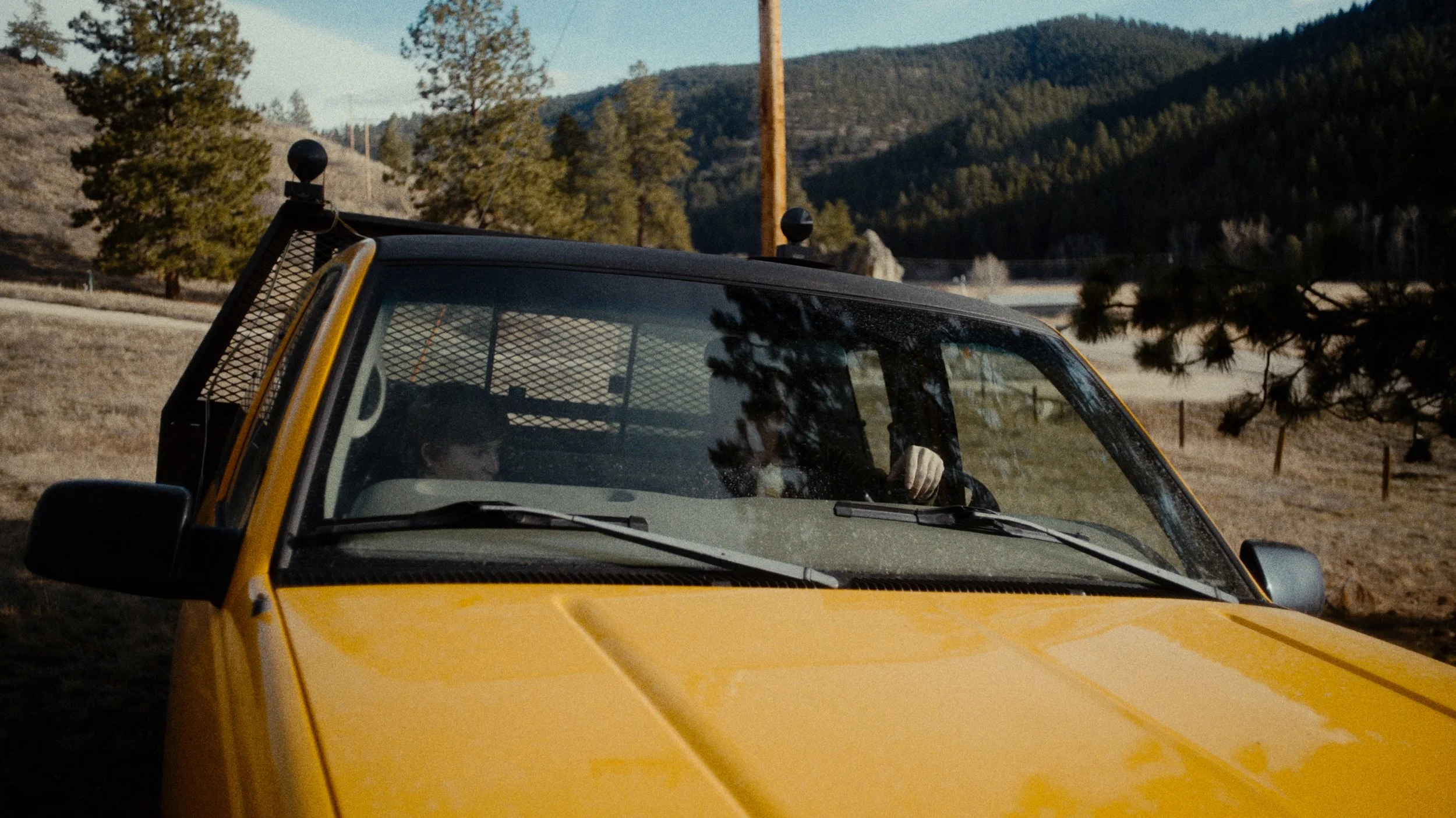 A yellow utility vehicle with two people inside, one in the driver's seat and one in the passenger seat, parked outdoors near trees and mountains.