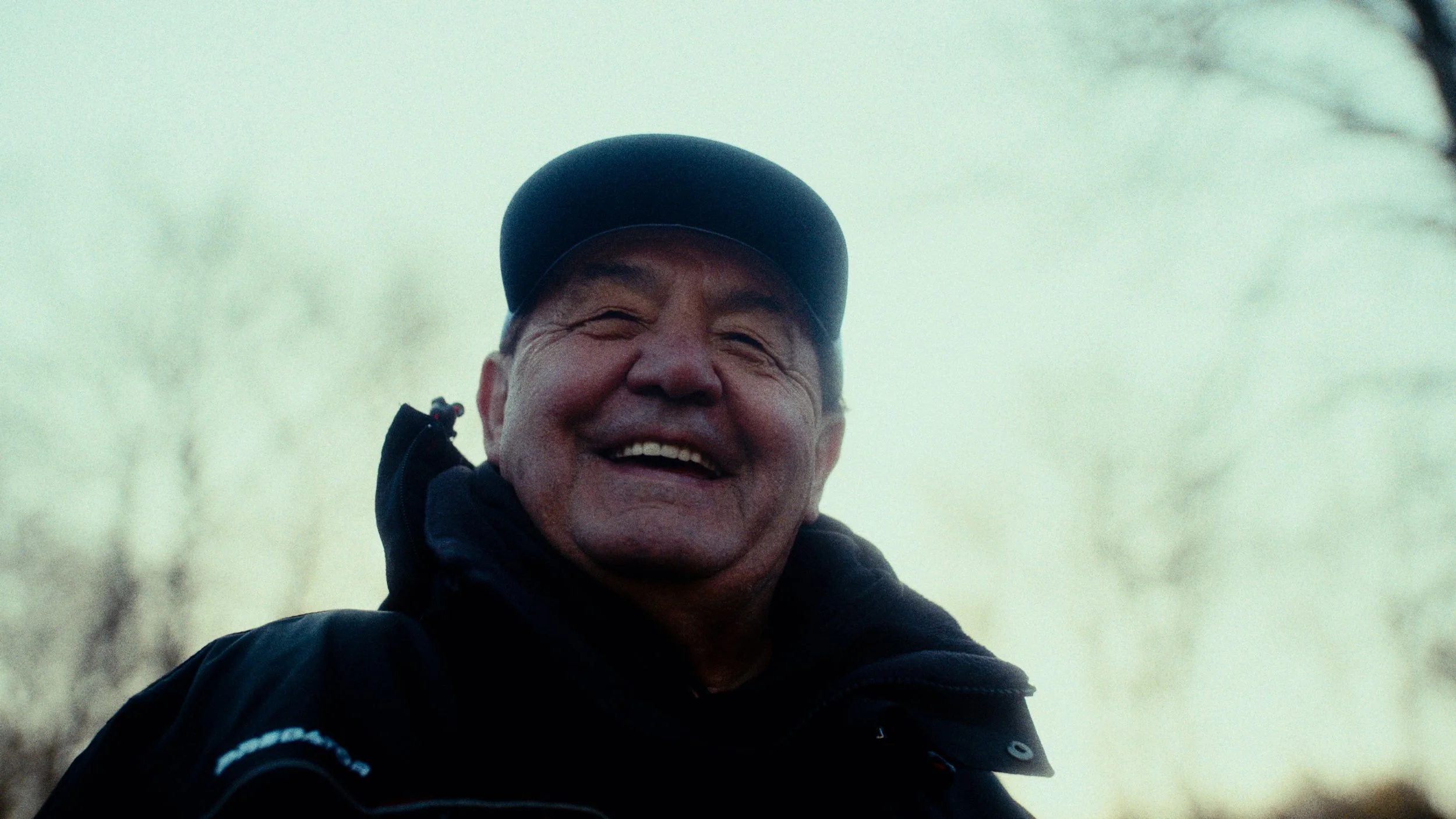 A middle-aged man outdoors, smiling and wearing a black cap, black jacket, and gloves, with a blurred background of trees.