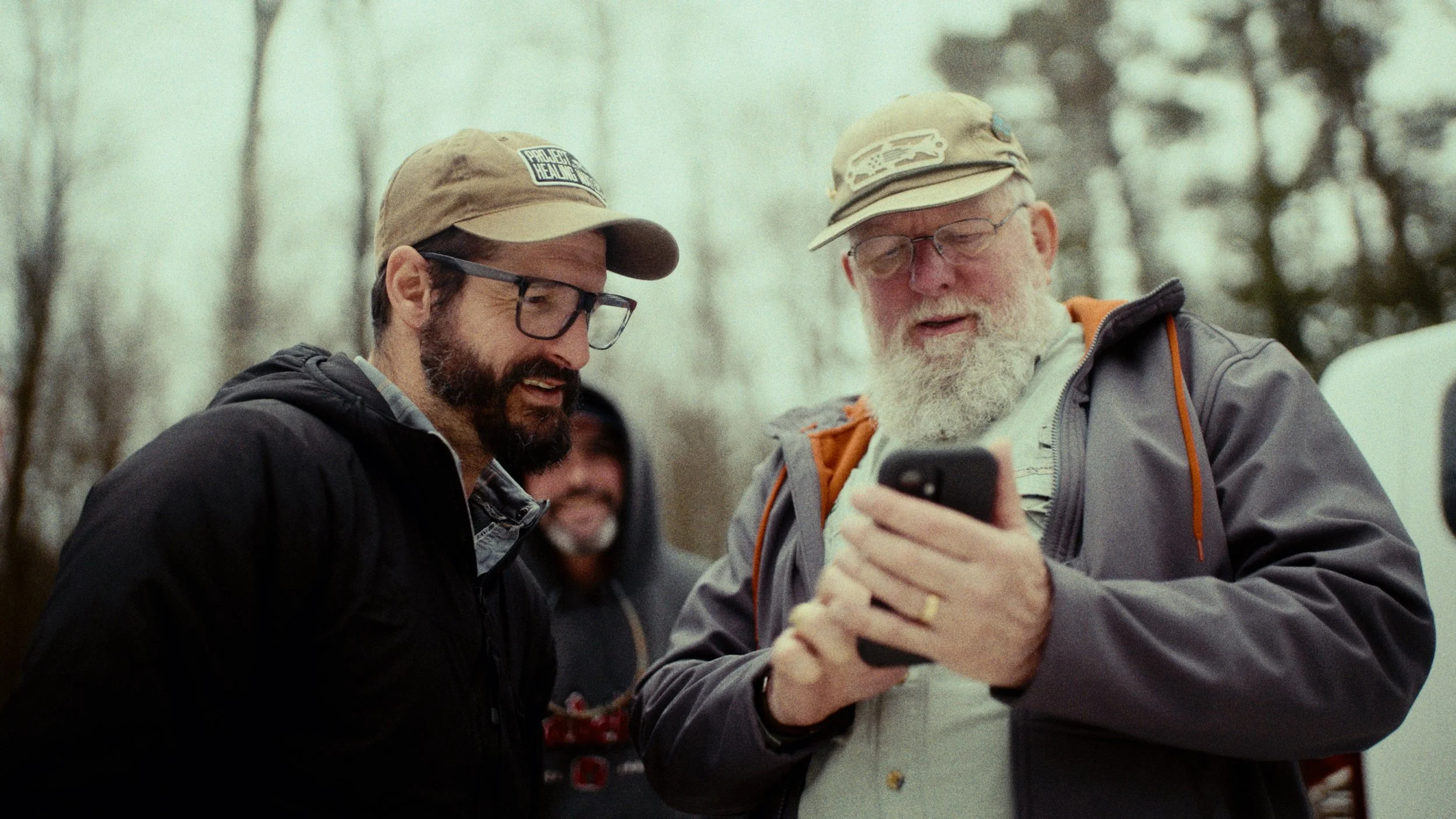 Two men looking at a smartphone together outdoors, trees in the background, one with glasses and a beard, wearing a cap and jacket, the other with a white beard and glasses, wearing a cap and jacket.