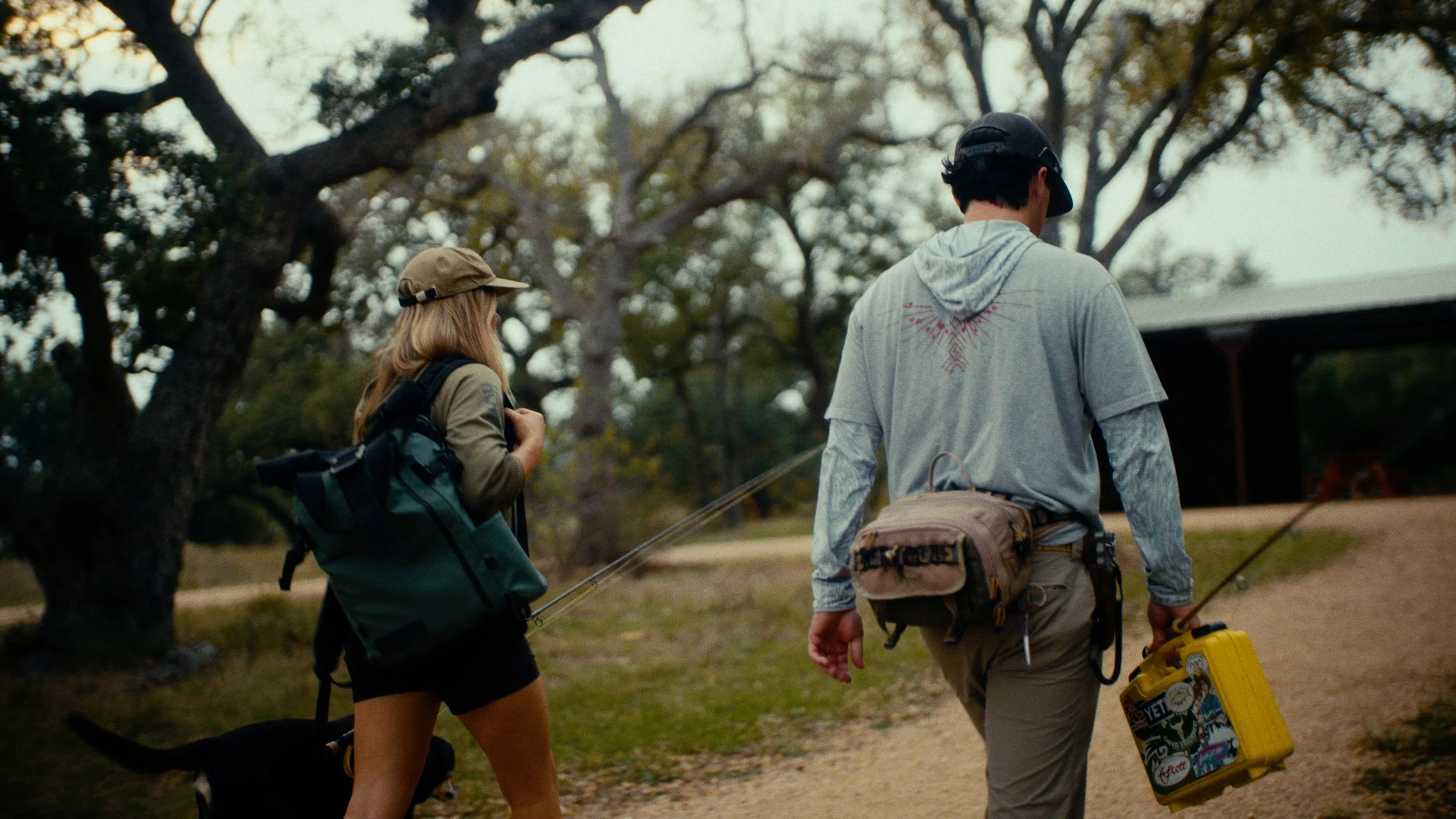 Two people, a woman and a man, walking on a dirt path in a park or wooded area. They are carrying backpacks and hiking or camping gear, with the man holding a yellow suitcase. The woman has a small dog on a leash. Trees surround the path, and a build