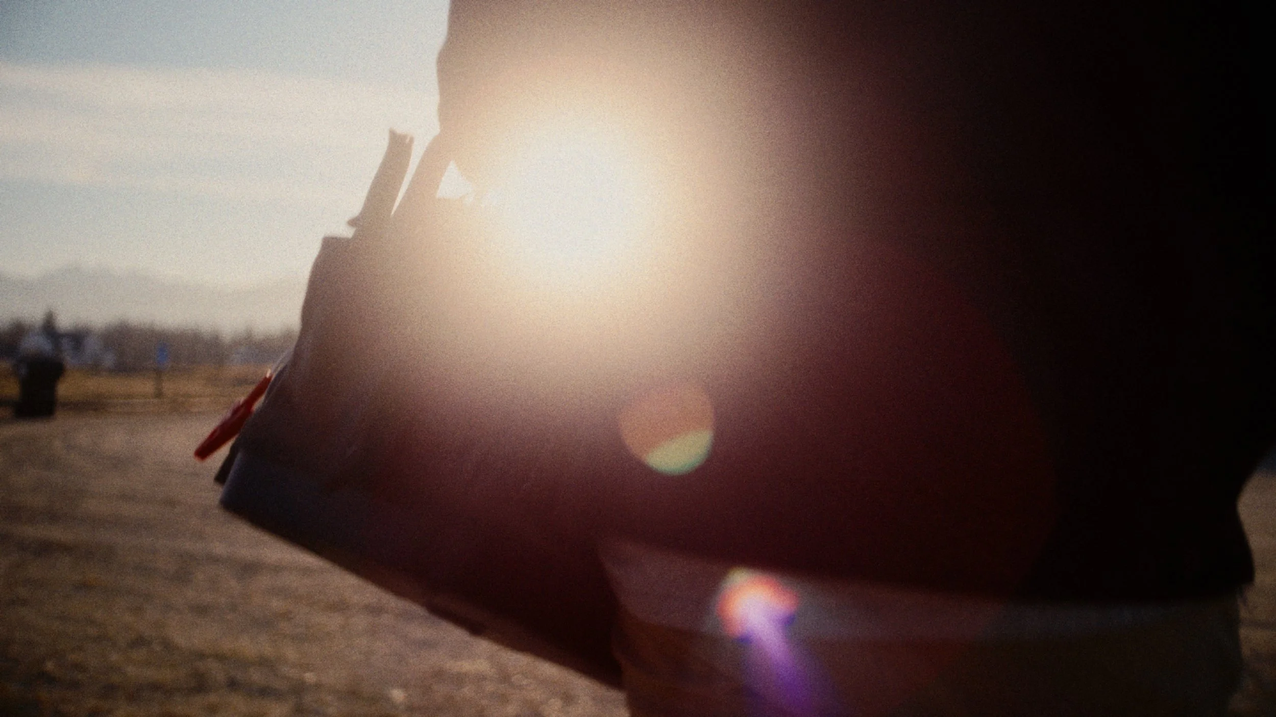 A person holding a book or magazine in front of the sun, creating a lens flare effect, with a distant landscape and mountains in the background.