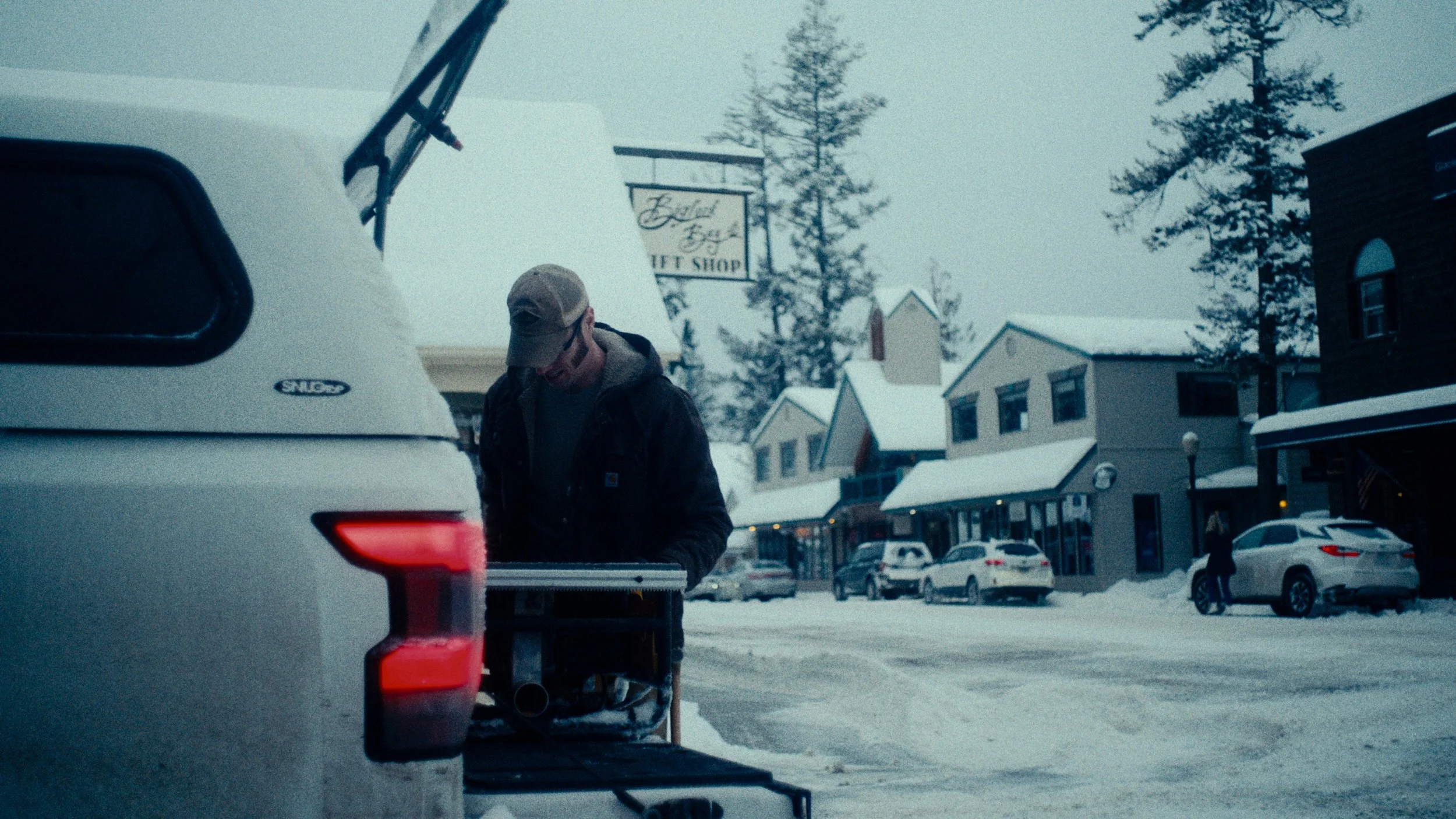 A man in a gray cap and dark jacket working outdoors in a snowy town, with parked cars and buildings in the background.