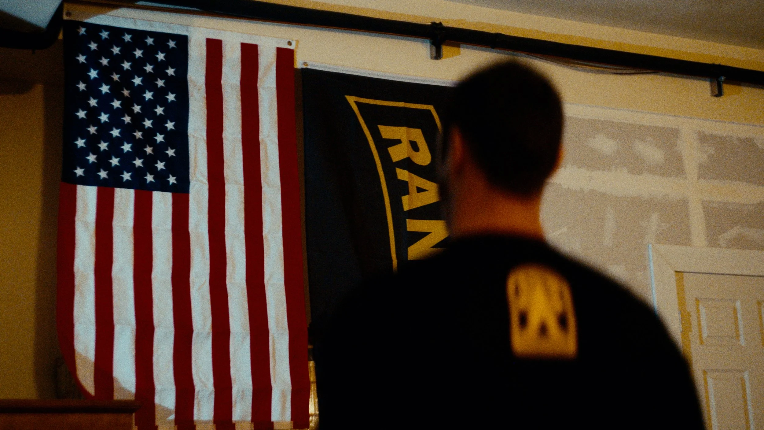 A person in a black jacket with a yellow patch on the shoulder stands in front of an American flag and a RAVN banner hanging on the wall.