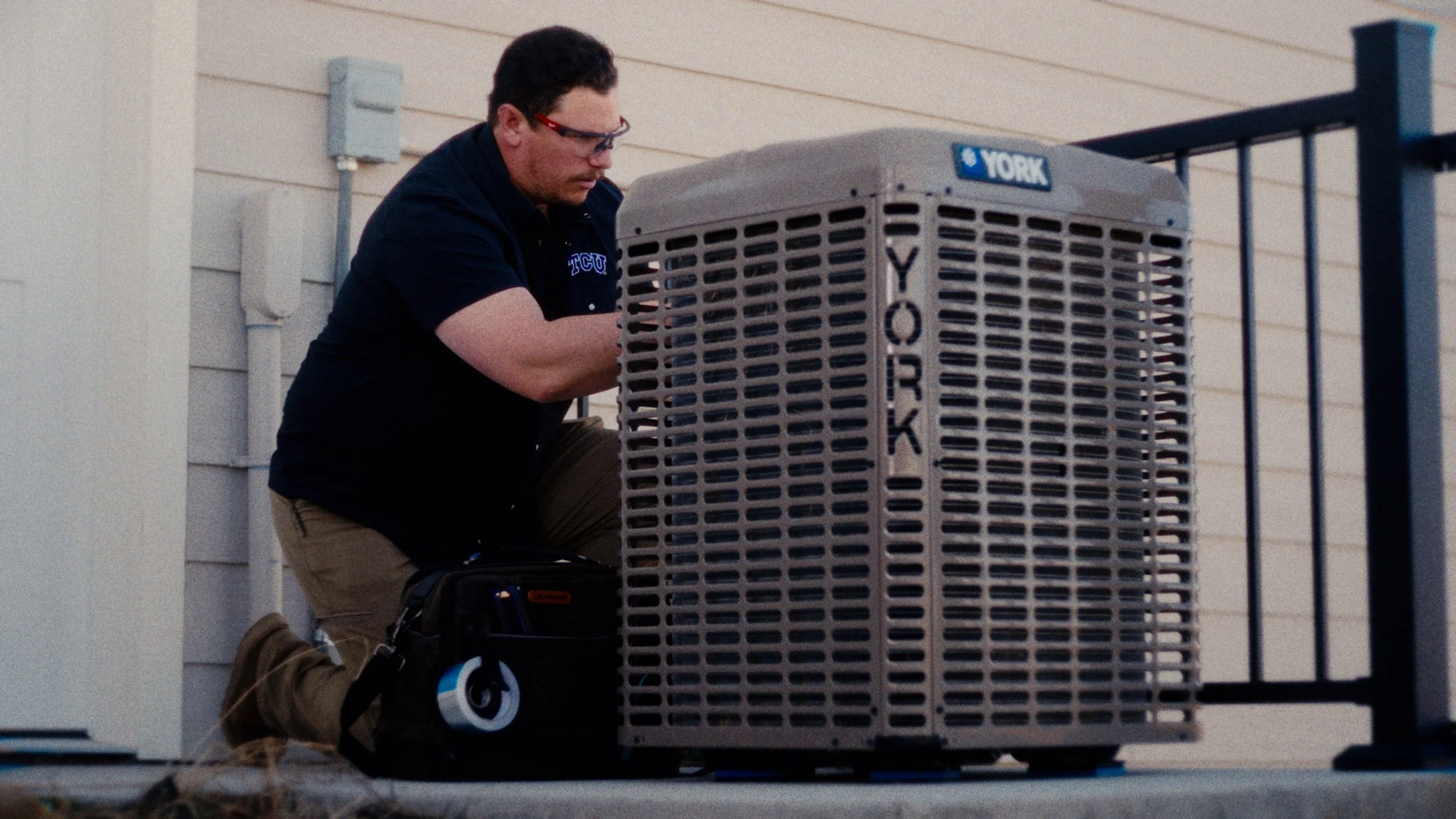 A technician kneeling on a porch, working on an outdoor York air conditioning unit next to a house wall with beige siding.