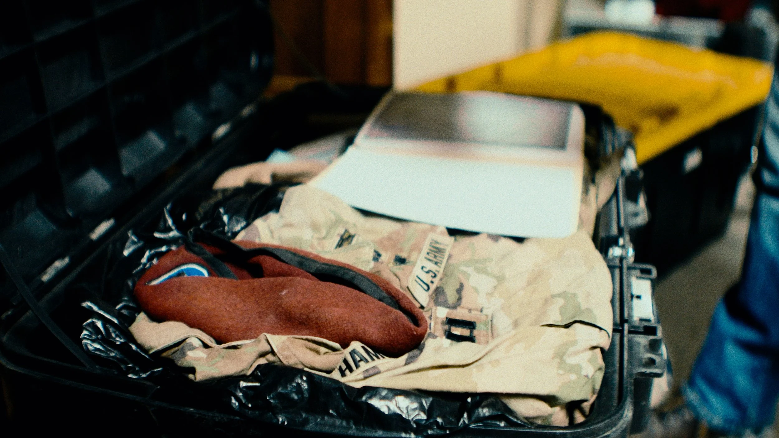 A military camouflage jacket, a red patch, and a small checkered pouch inside an open black suitcase