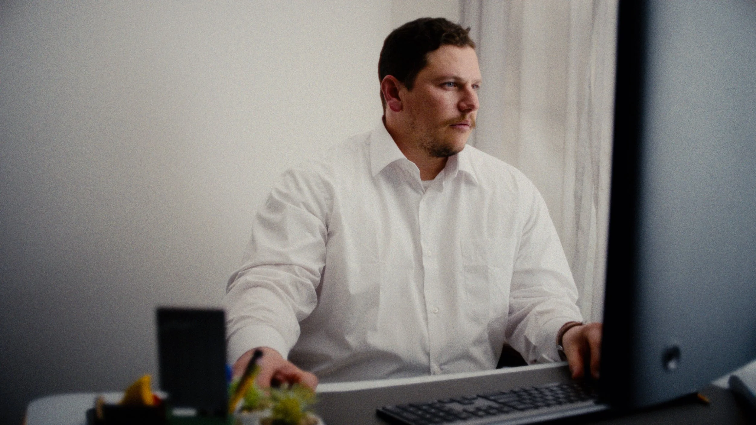 A man in a white shirt working on a computer at a desk, with a blurred foreground and a plain light-colored wall in the background.