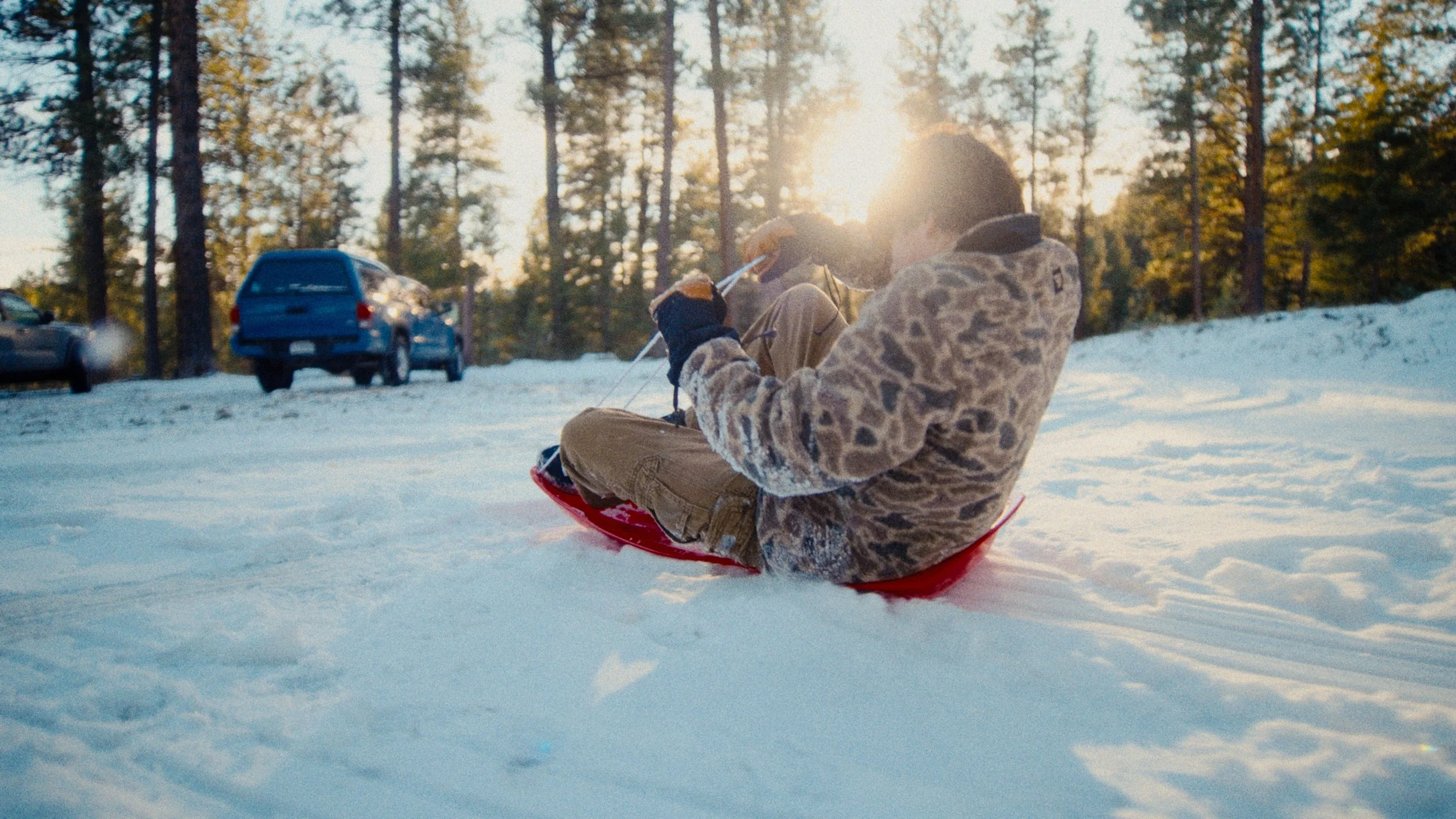 A person sitting on a red sled in a snowy landscape, with cars parked nearby and trees in the background, during sunset.