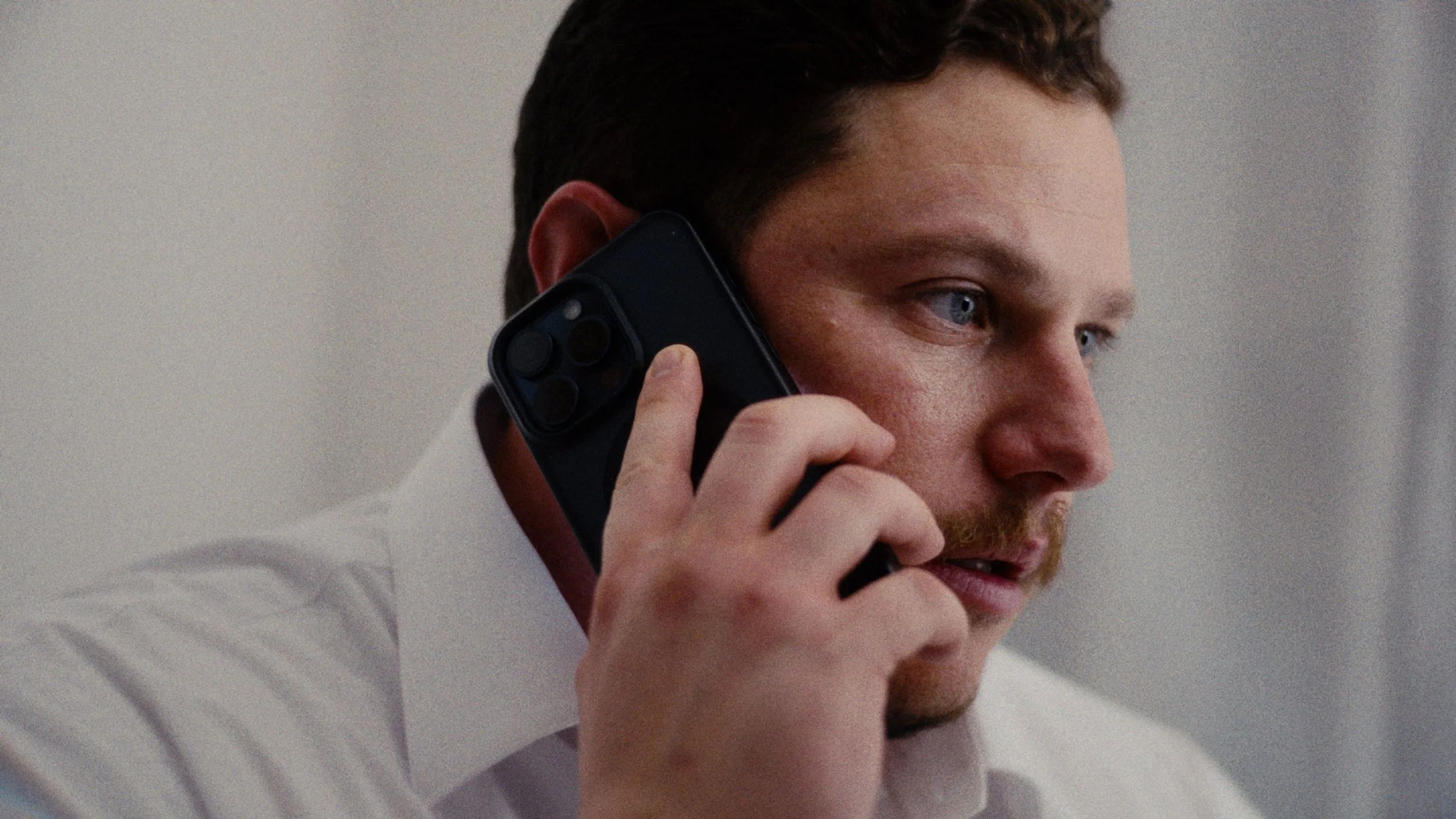 A man with short brown hair and a beard, wearing a white shirt, speaking on a black smartphone with a triple camera setup, indoors with a light-colored wall in the background.