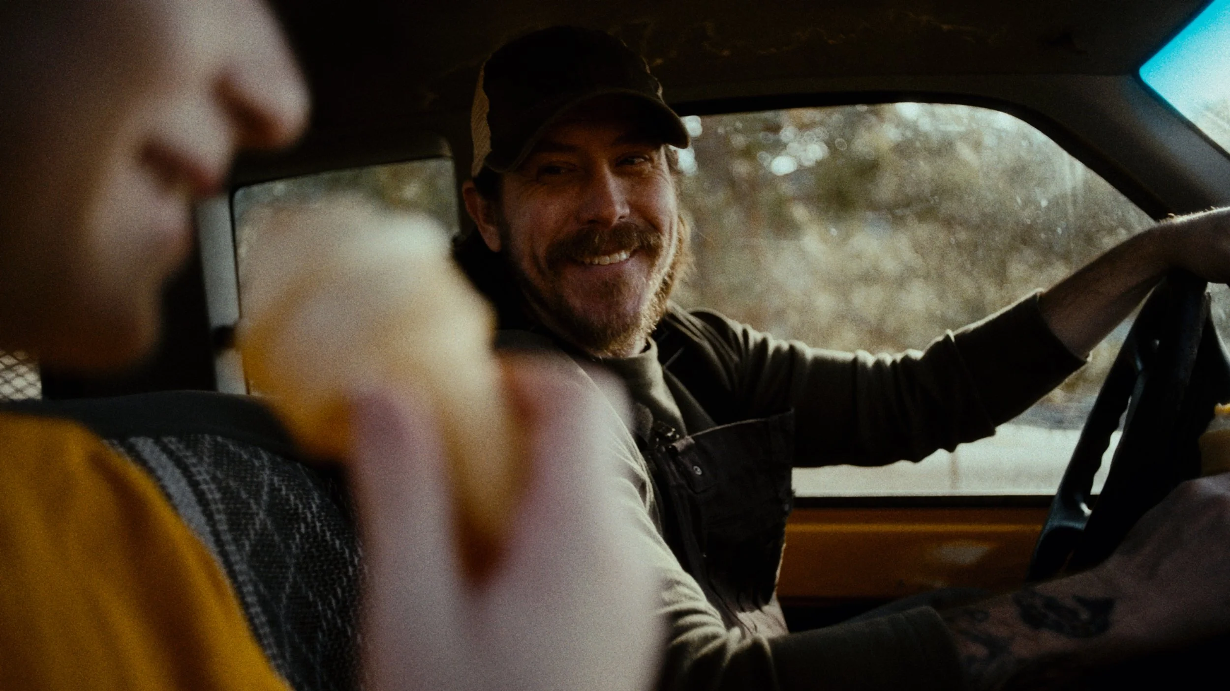 A man driving a vehicle and smiling at someone, with a person in the passenger seat eating an ice cream cone.