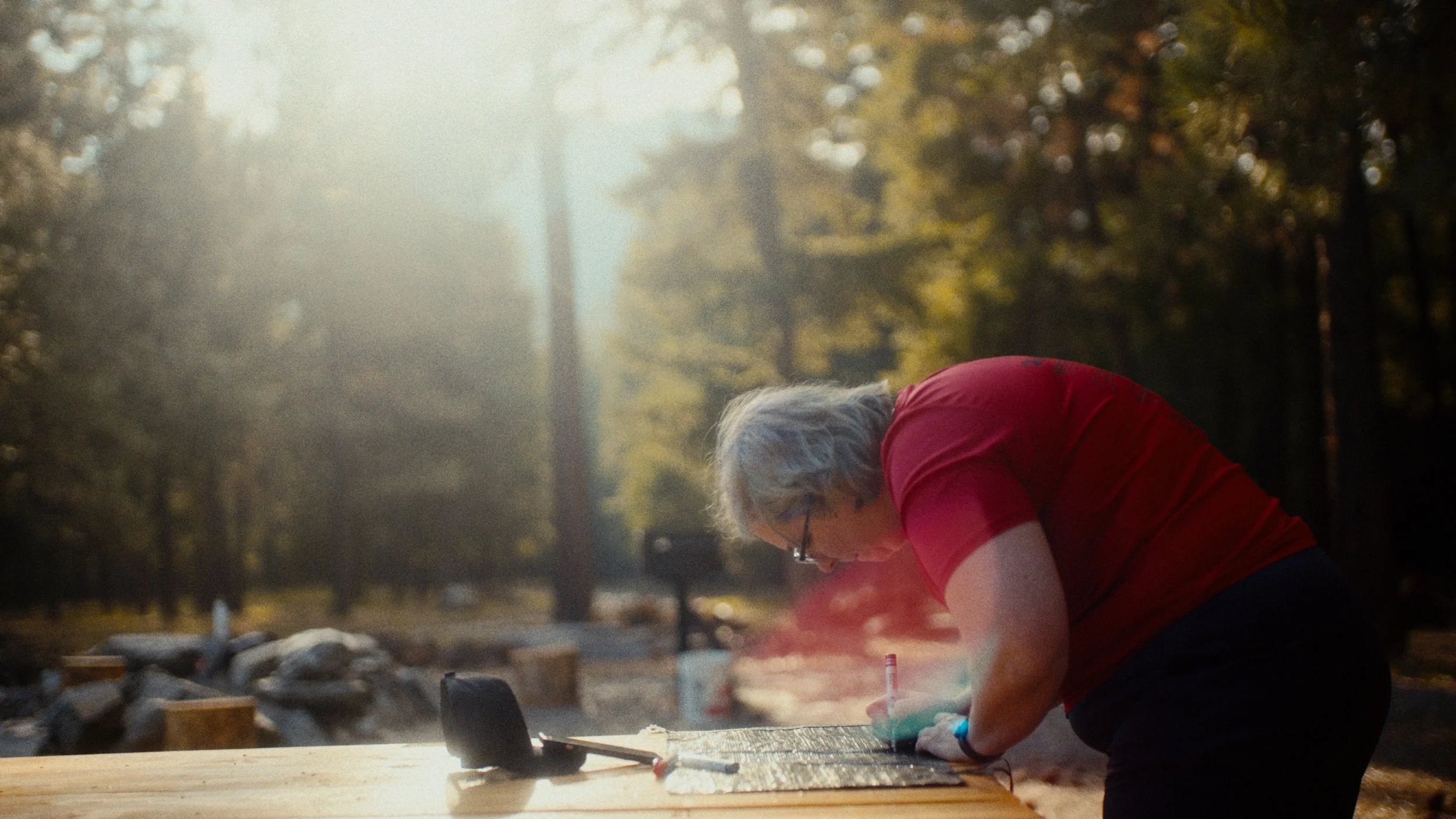 Person with gray hair and red shirt writing on a board outdoors during sunset, with trees and a forest in the background.