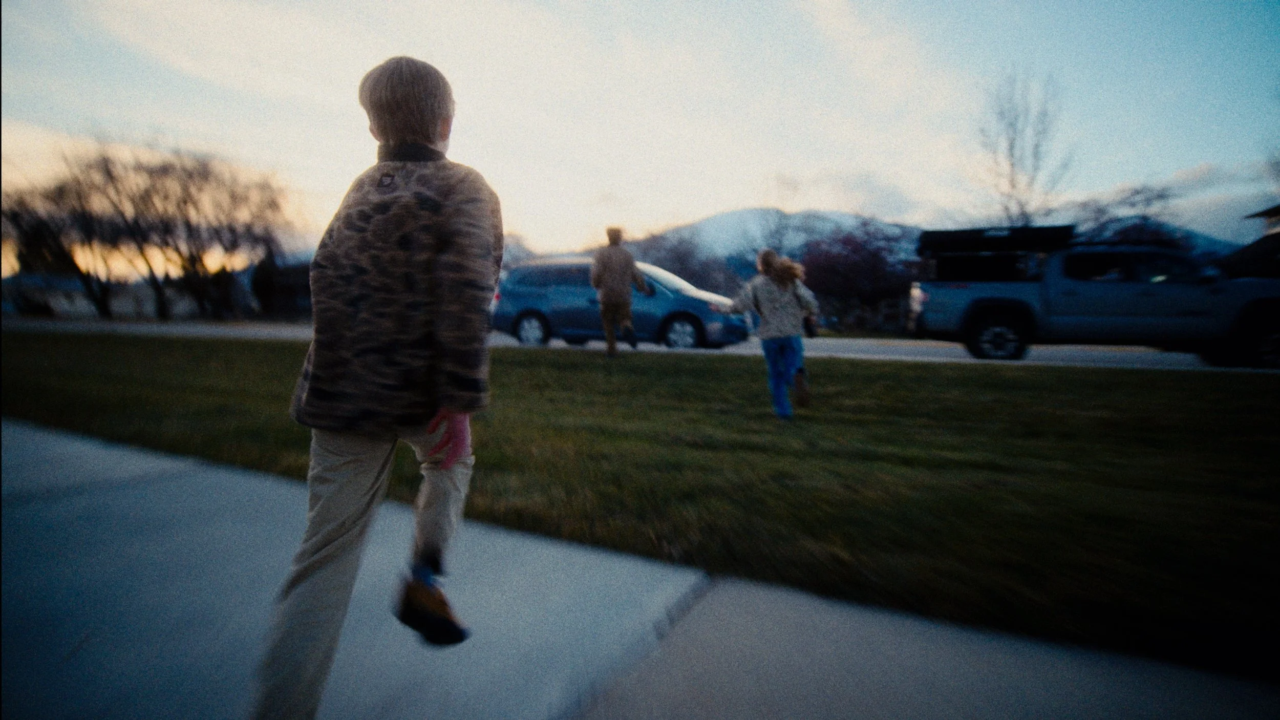 A woman with children running outdoors on a sidewalk during sunset, with mountains, trees, and parked cars in the background.