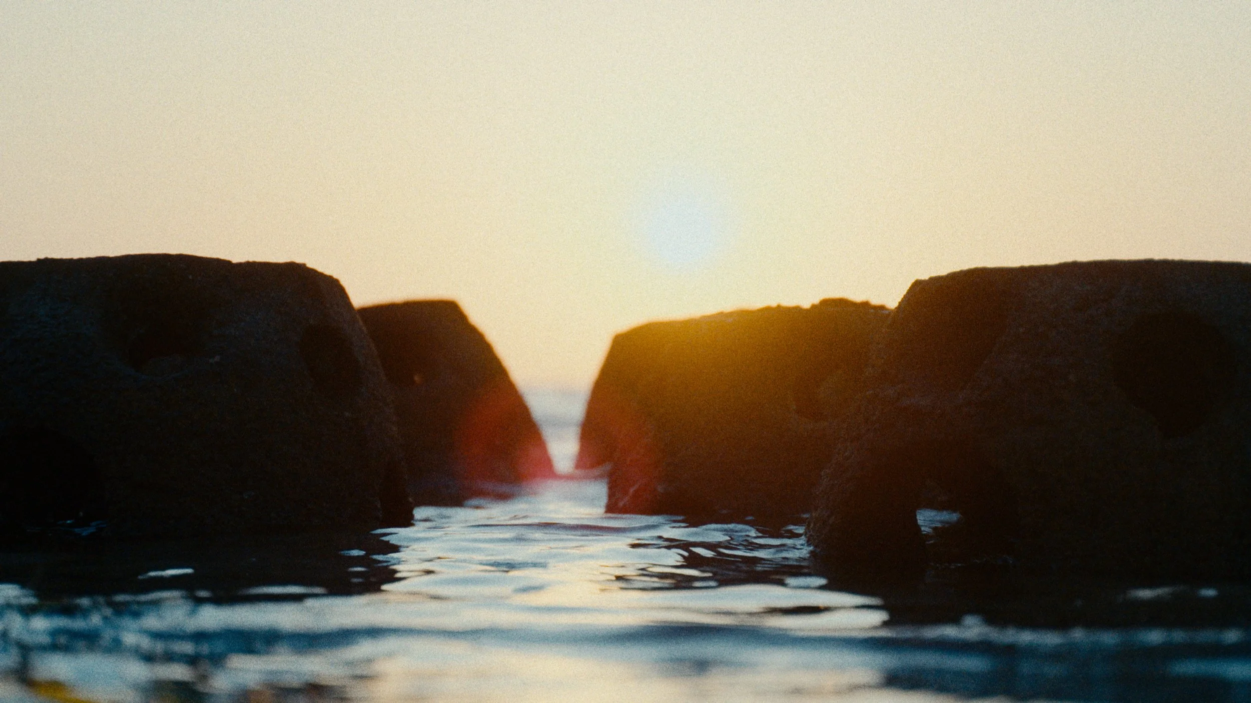 Sunset viewing between two large dark rocks in the ocean with gentle waves, warm glowing sky.