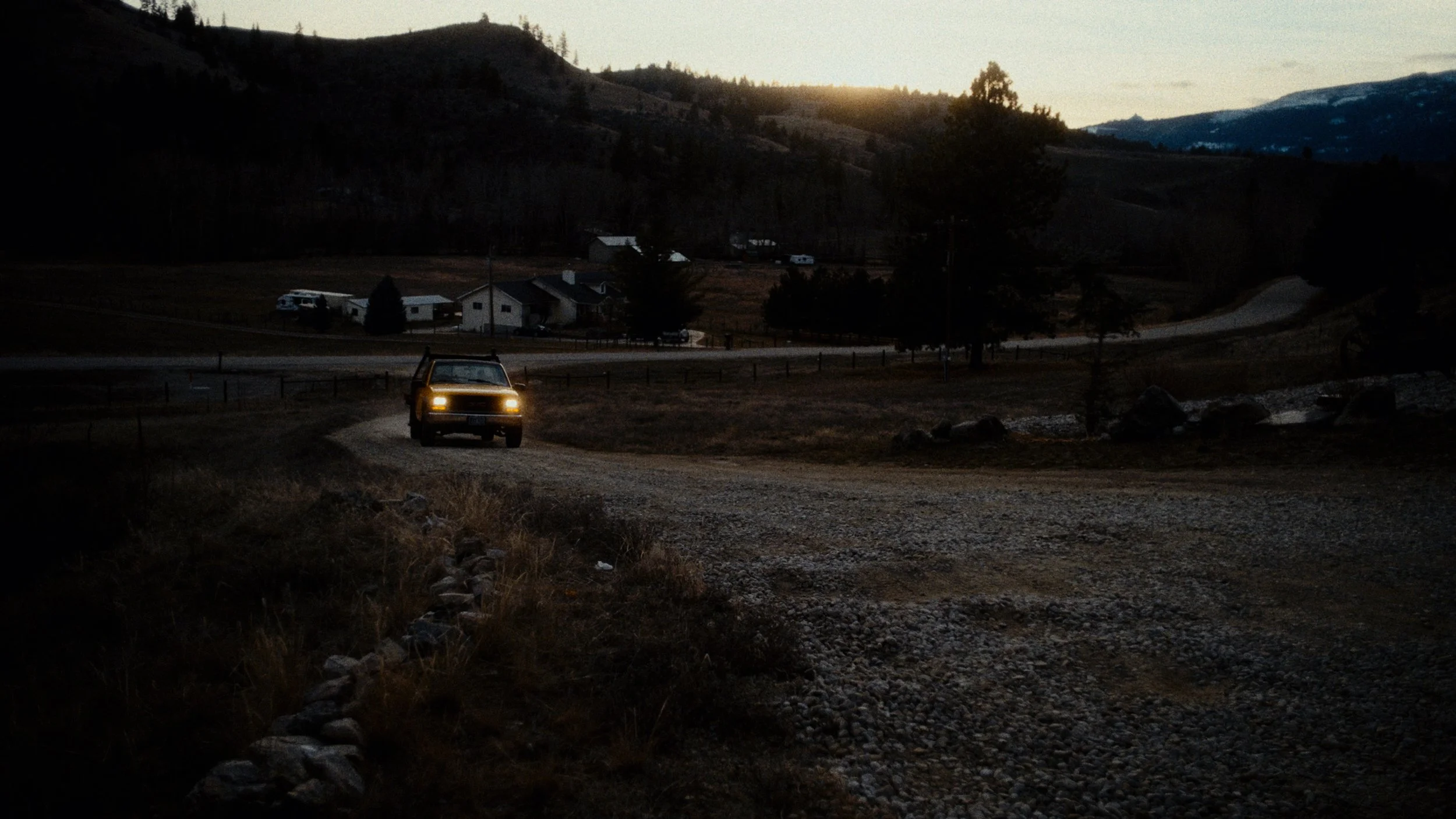 A vehicle drives down a dirt road in a rural landscape at dusk, with houses and trees on rolling hills in the background under a setting sun.