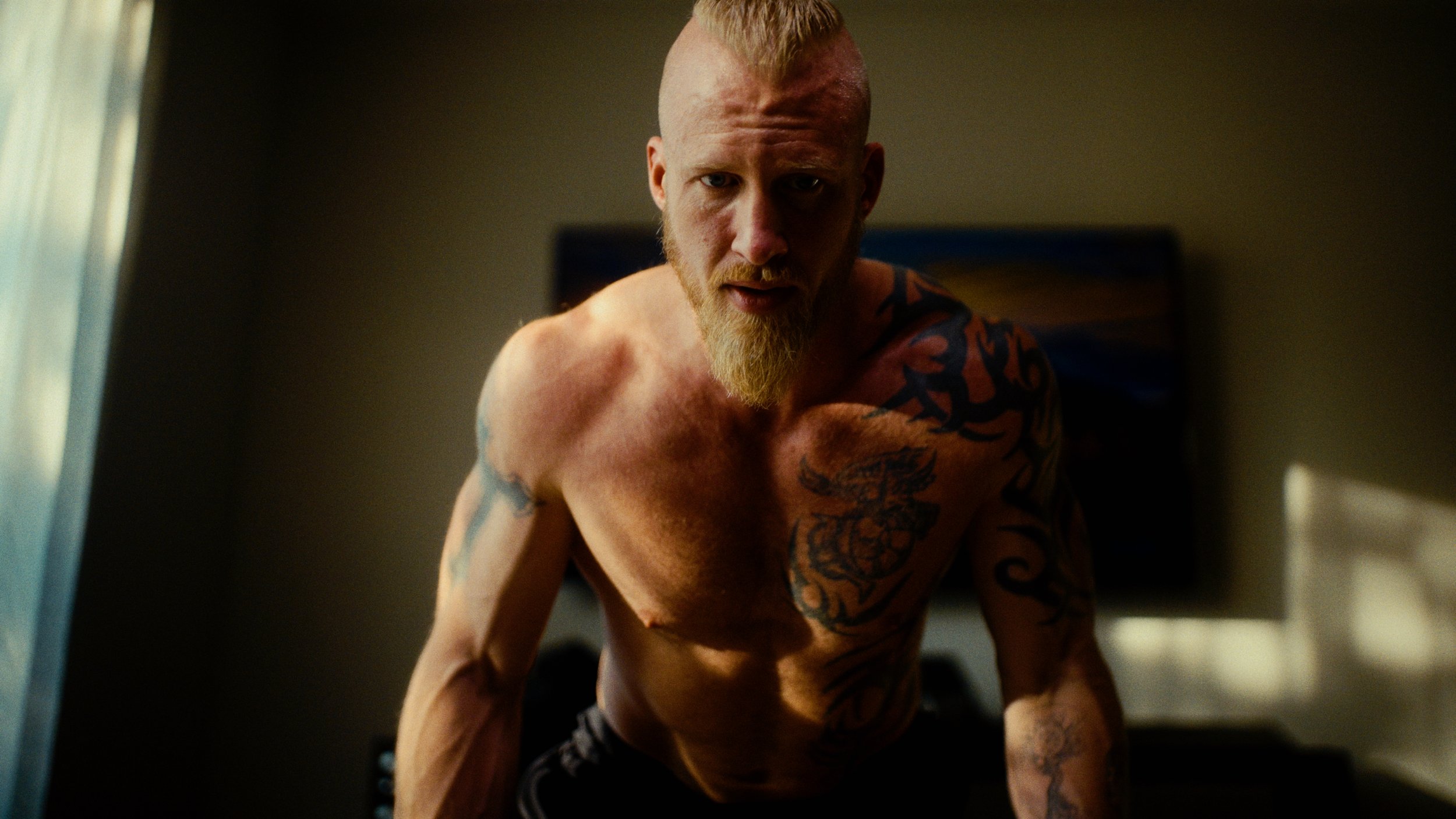 A shirtless man with tattoos on his chest and arms, with a mohawk hairstyle, looks directly into the camera in a dimly lit room.